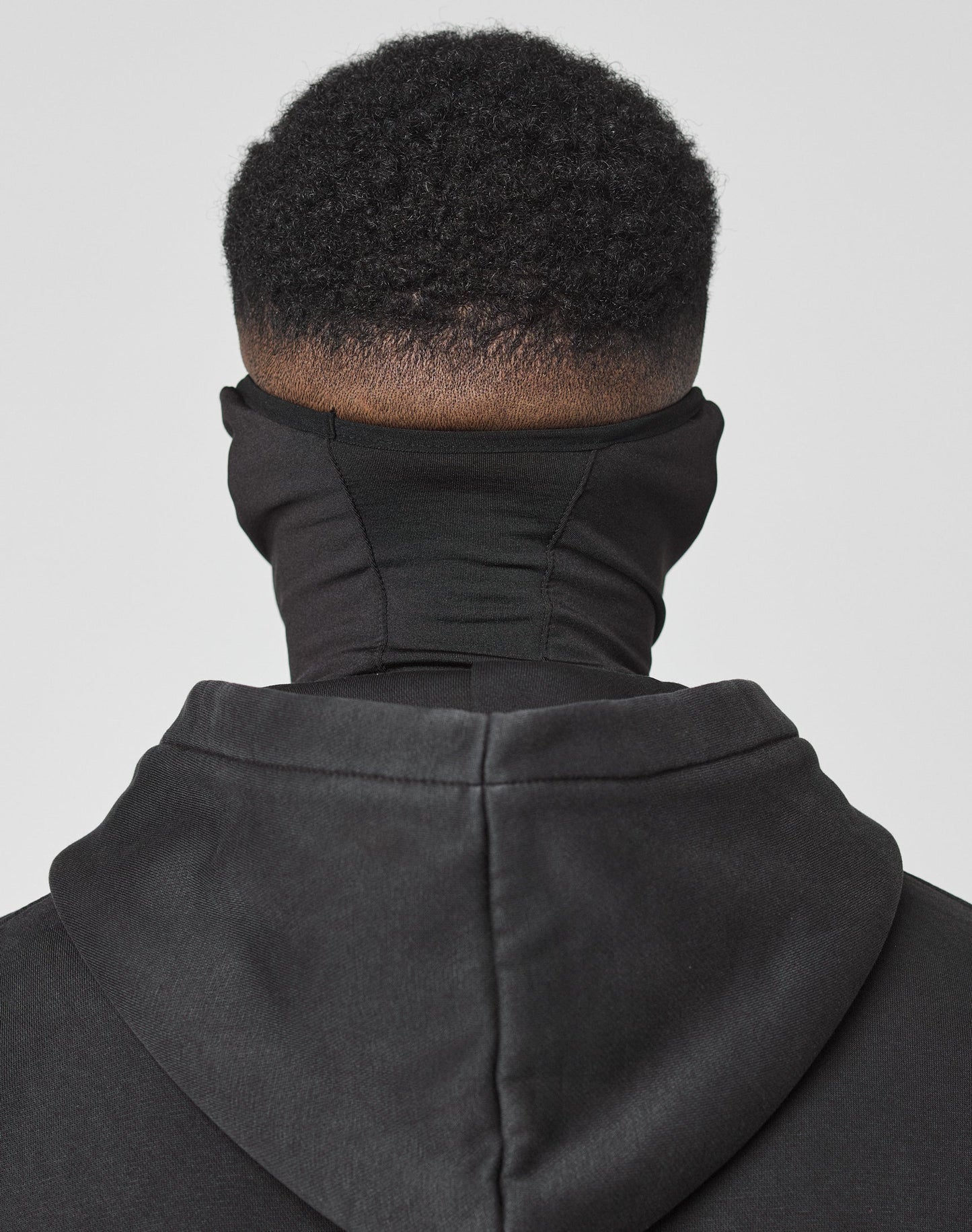 A person with short, curly hair, seen from behind, wears a black 100% polyester LIVE FAST DIE YOUNG hoodie with reflektierender Druck and the Live Fast Performance Face Mask. The background is plain light gray; Waschhinweise are not visible.