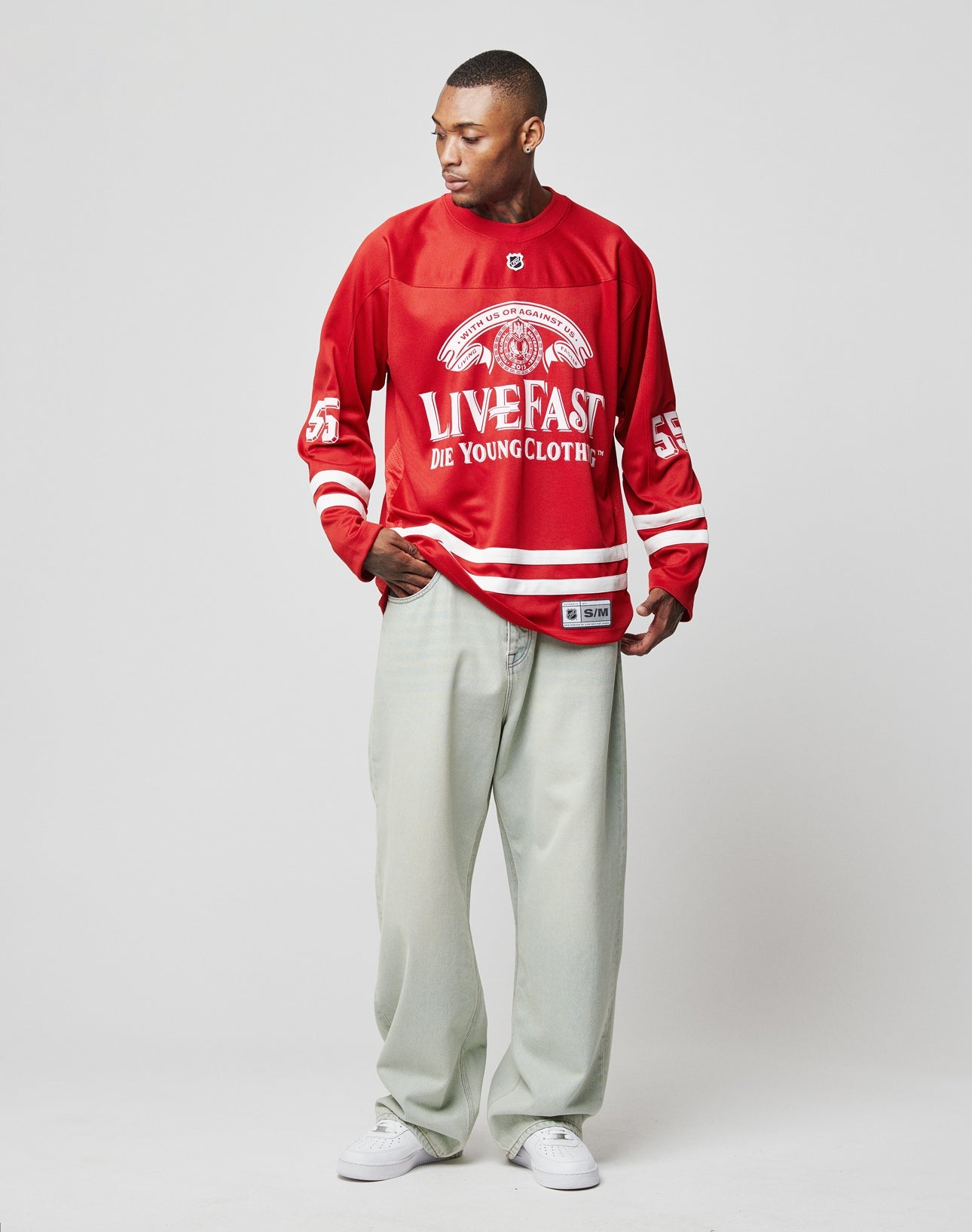 A man stands against a plain background in an oversize-fit red LFDY Hockey Jersey by LIVE FAST DIE YOUNG, paired with light gray wide-leg pants and white sneakers. He looks to his left, one hand holding his shirt.