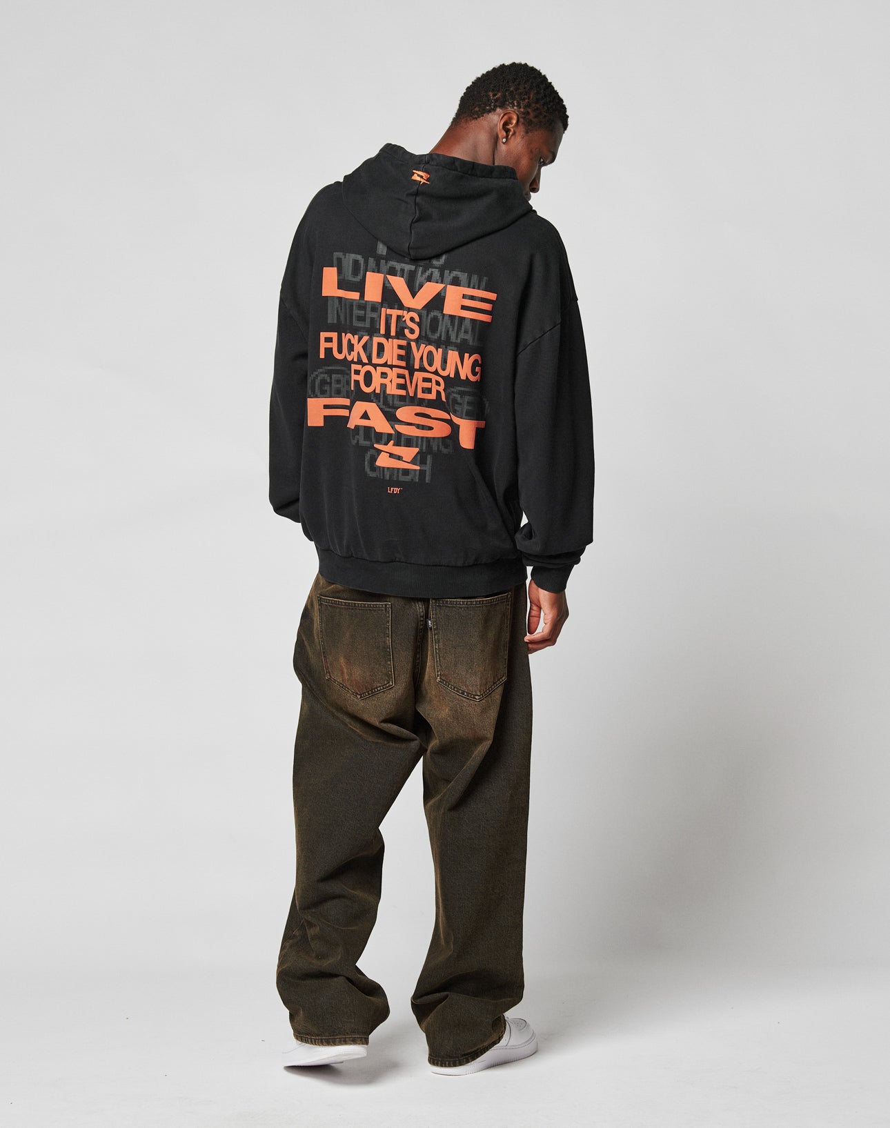 A man stands facing away, wearing the LIVE FAST DIE YOUNG Print Over Hooded in black with bold orange text and baggy dark jeans. The oversize fit hoodie reads, “LIVE INH, IT’S OVA...” on a light gray background.