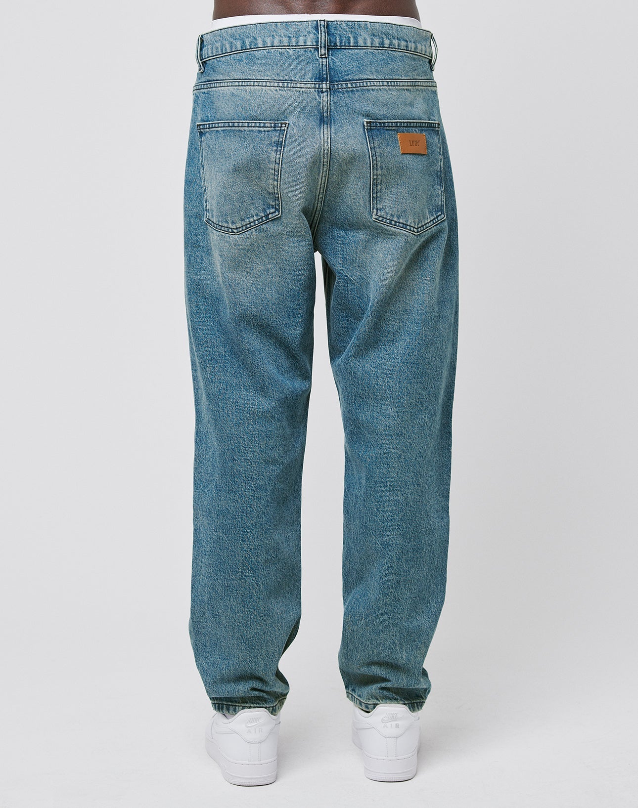 A person is seen from the back wearing LIVE FAST DIE YOUNG Regular Denim blue jeans with a tapered leg, two rear pockets, and a brown leather patch on the right pocket, paired with white sneakers against a white background.
