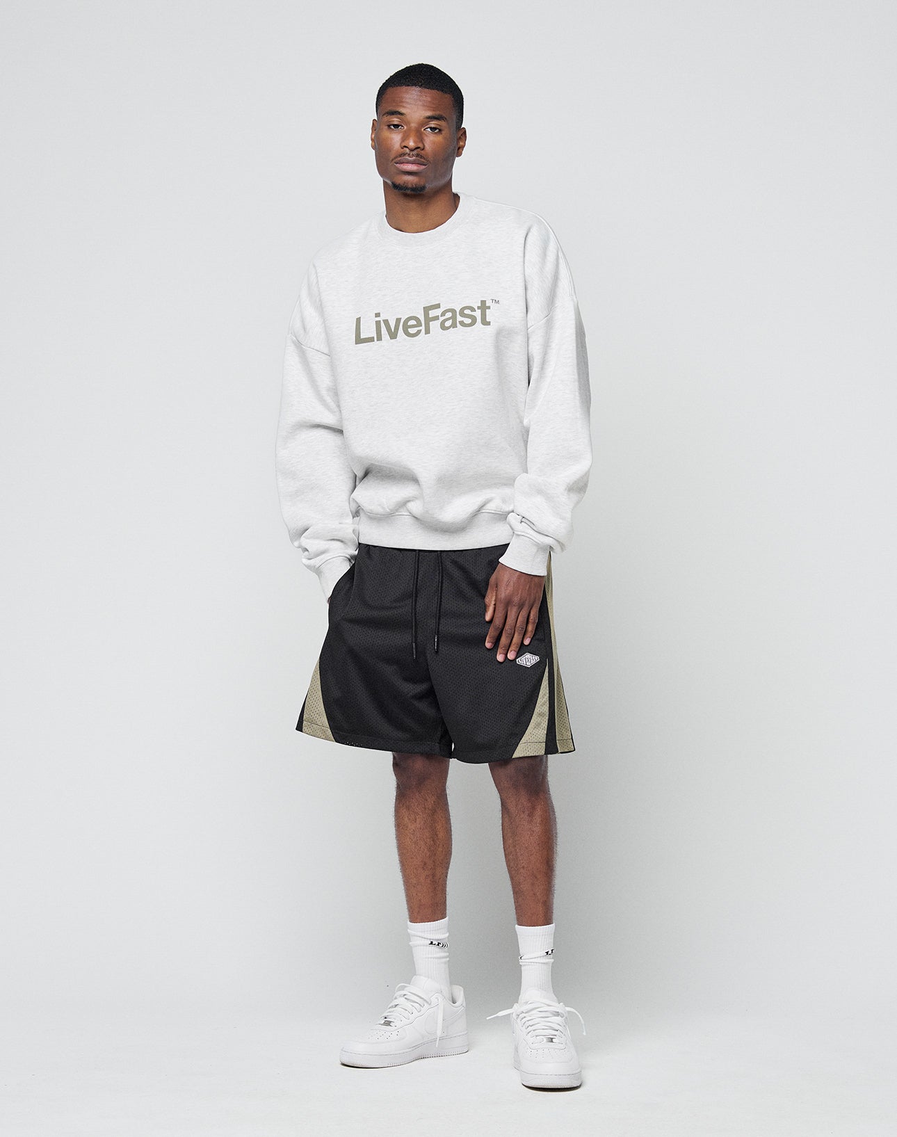 A man stands against a plain light background wearing the Basic LiveFast Sweater by LIVE FAST DIE YOUNG, a gray oversize fit with LiveFast print, styled with black shorts with olive sides, white socks, and sneakers for a comfy-cool look.