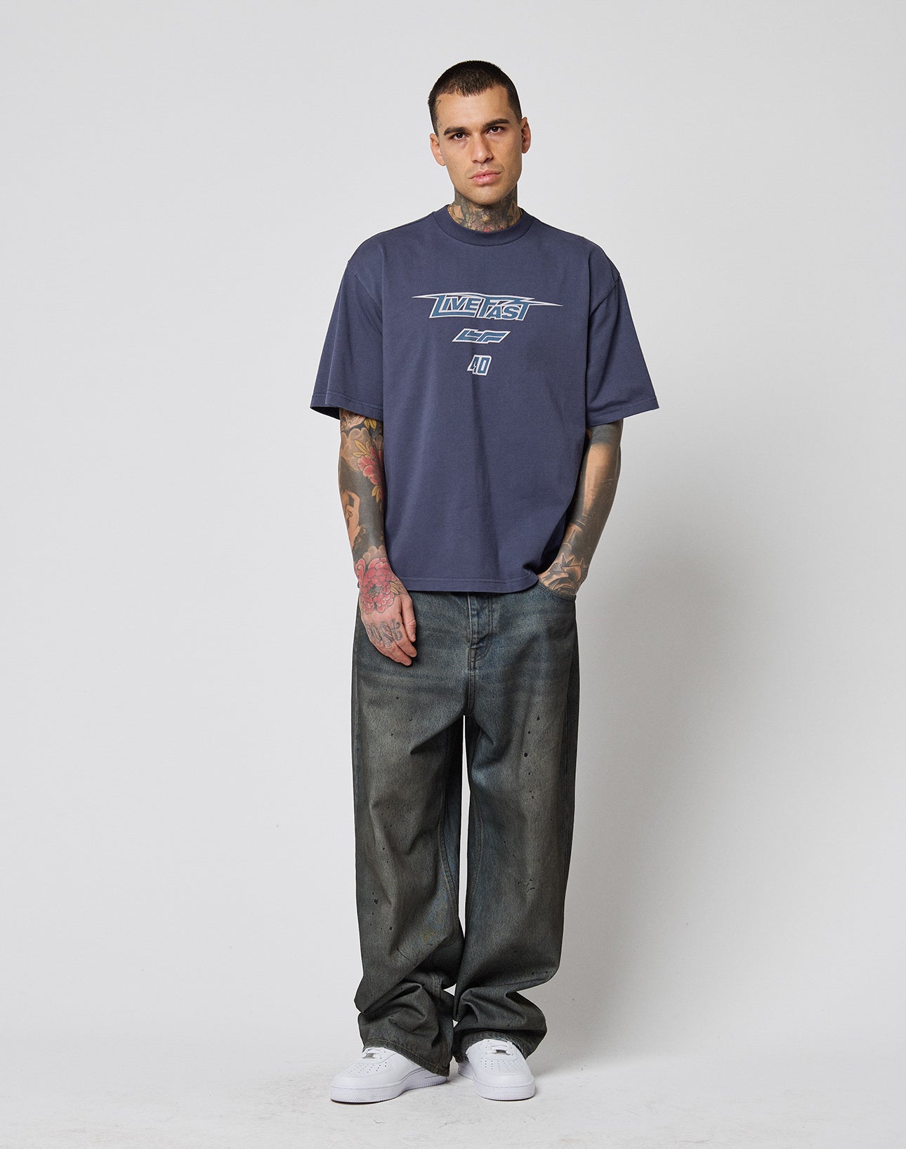 A person with short hair and visible tattoos stands against a plain white background, wearing an oversized dark blue LFDY Triple Racing Tee by LIVE FAST DIE YOUNG, paired with baggy faded jeans and white sneakers.