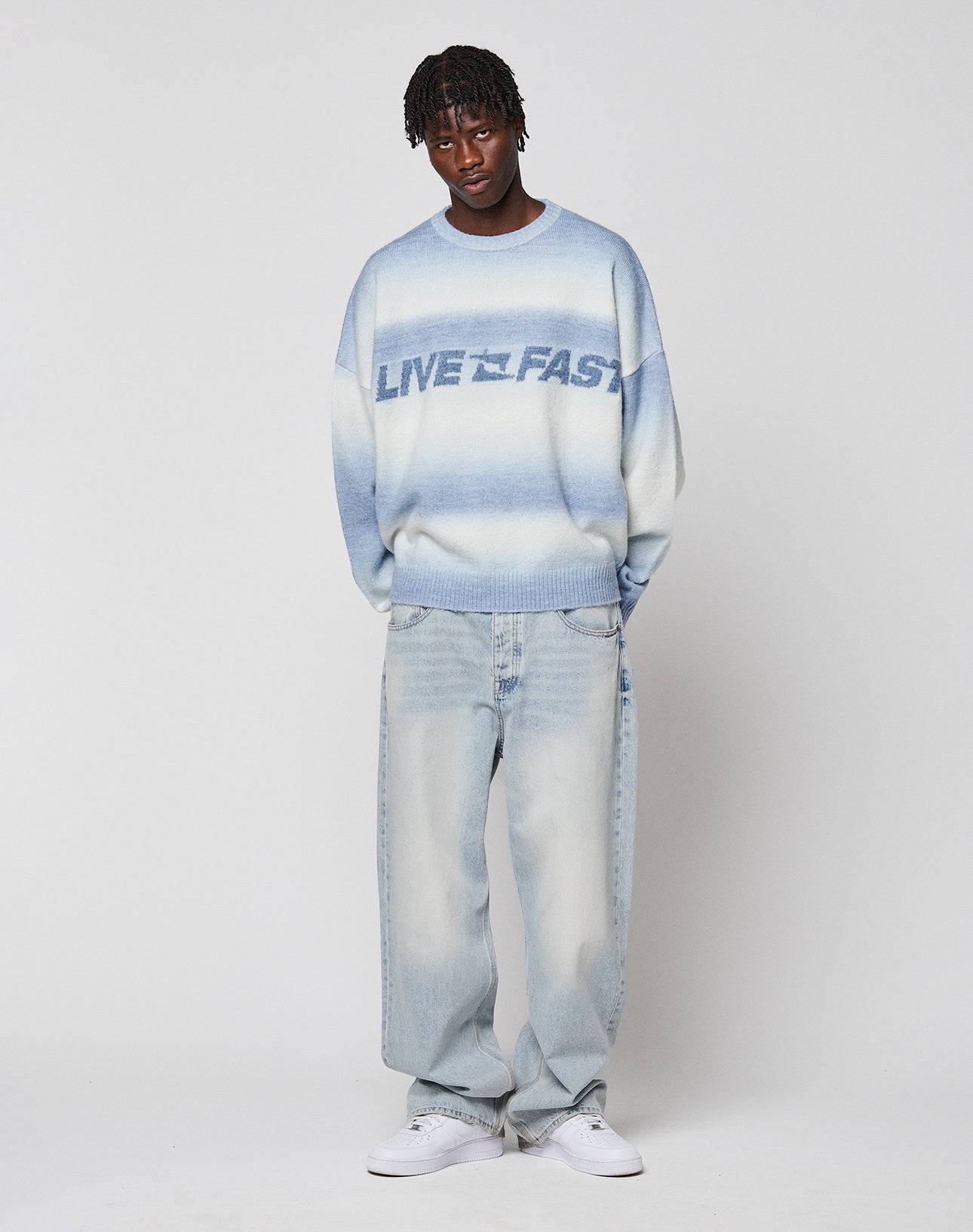 A person with short, braided hair wears the Live Fast Boucle Sweater by LIVE FAST DIE YOUNG in light blue and white, paired with loose faded jeans and white sneakers, standing against a plain white background.