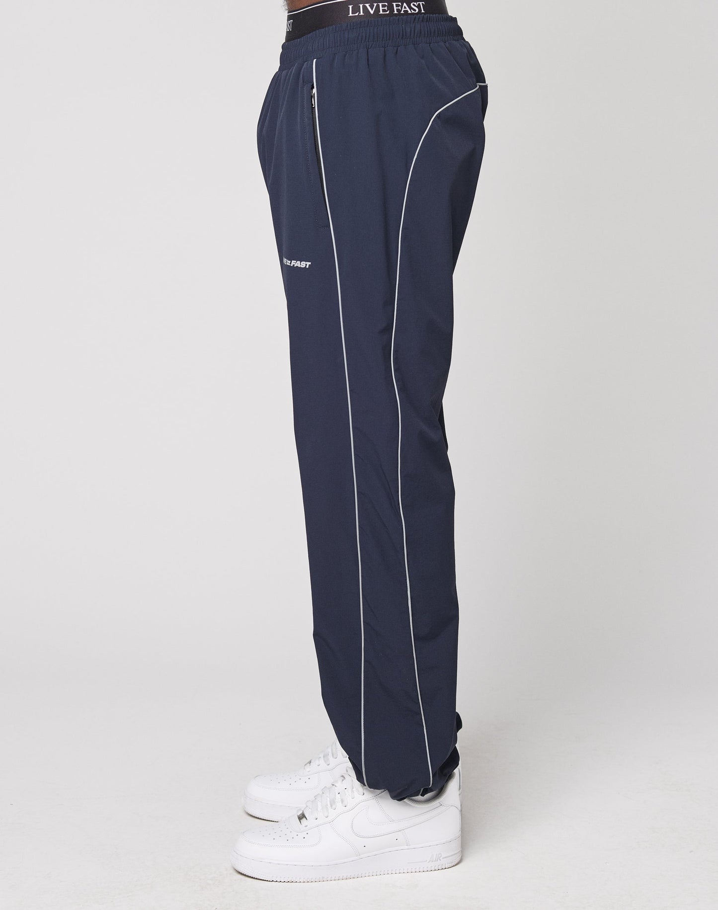 A model stands sideways against a white background wearing LIVE FAST DIE YOUNG’s navy Live Fast Trackpants with white piping, an elastic waistband featuring LIVE FAST in white text, and white sneakers.
