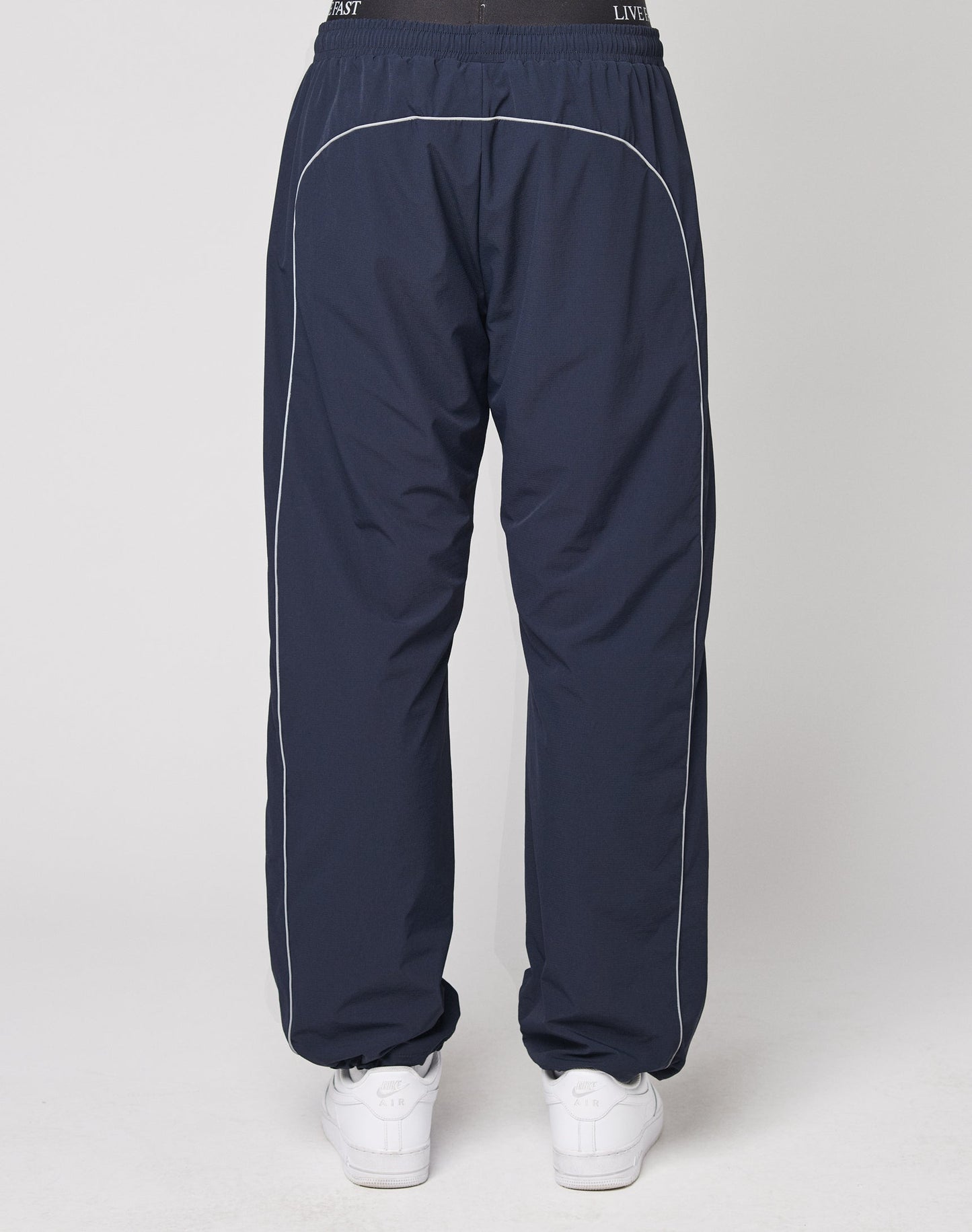 A person is seen from behind wearing LIVE FAST DIE YOUNGs Live Fast Trackpants in navy with white piping, a reflective logo print, and an elastic waistband in a wide fit, paired with white sneakers against a plain white background.