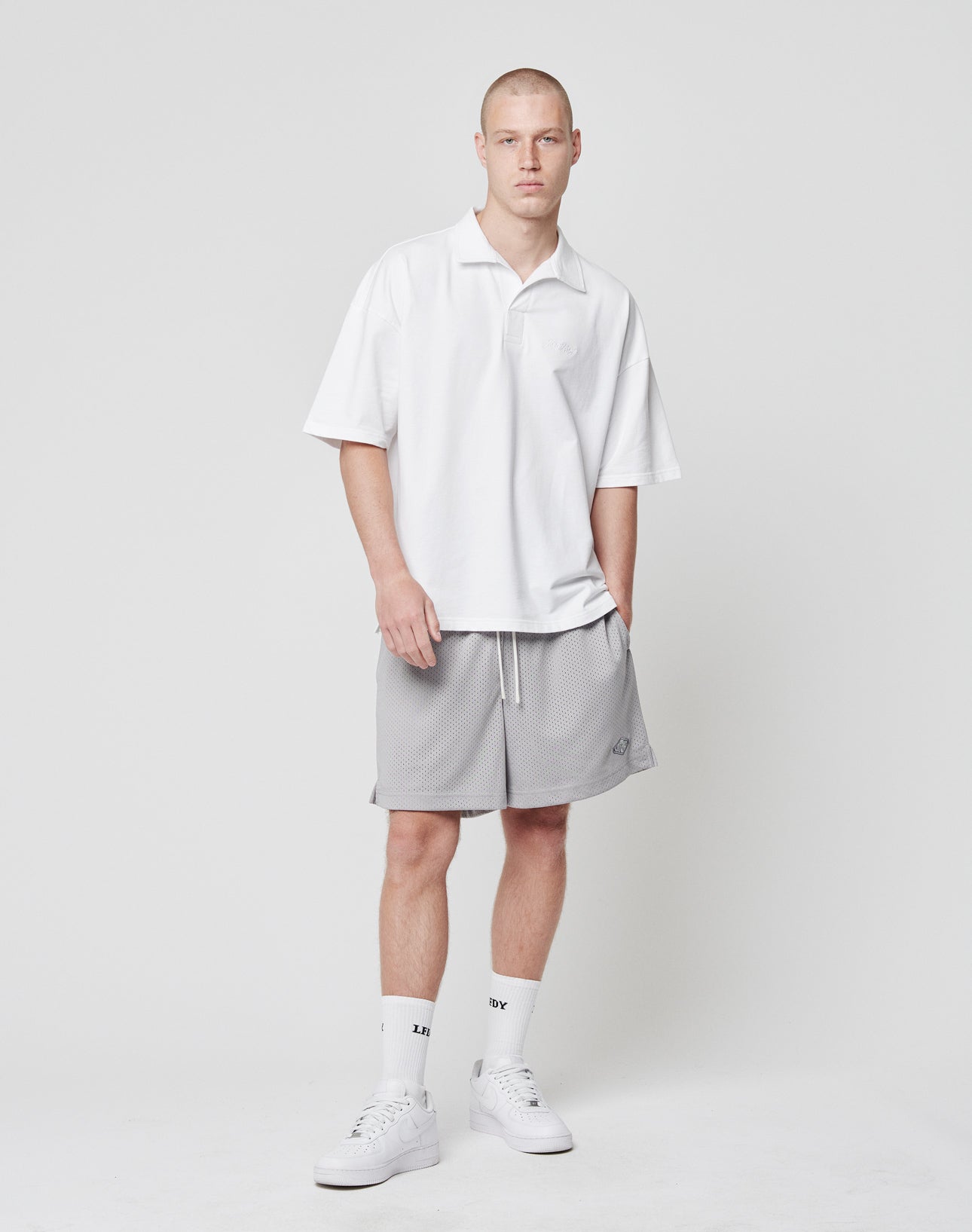 A young man with short hair and a neutral expression stands against a white background, wearing LIVE FAST DIE YOUNG Basic Mesh Shorts in light gray polyester, a loose white polo with logo patch, white crew socks, and sneakers.