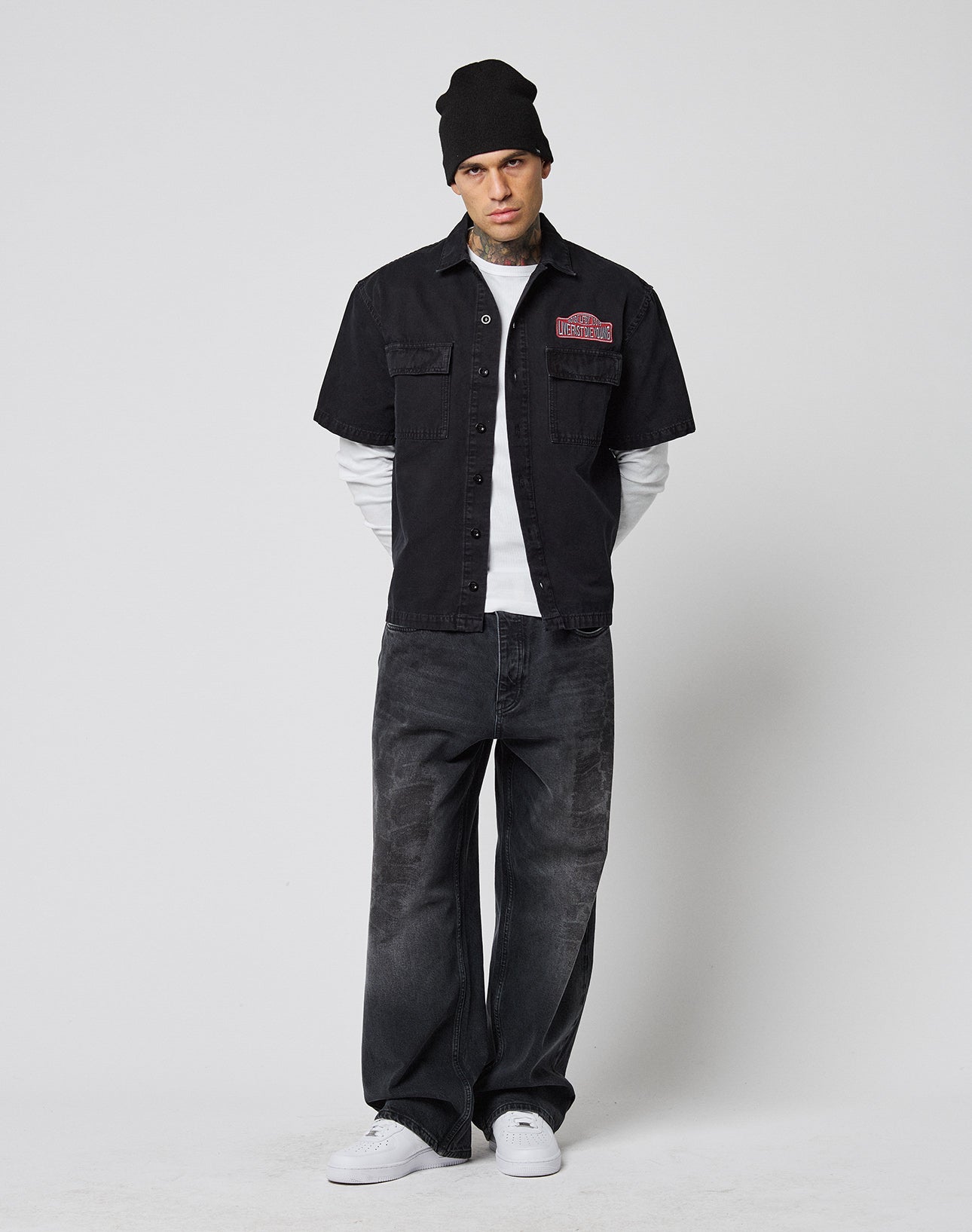 A person wears a black LFDY Workwear Shirt by LIVE FAST DIE YOUNG over a white long-sleeve, loose vintage black jeans, black beanie, and white sneakers, standing against a plain white background.