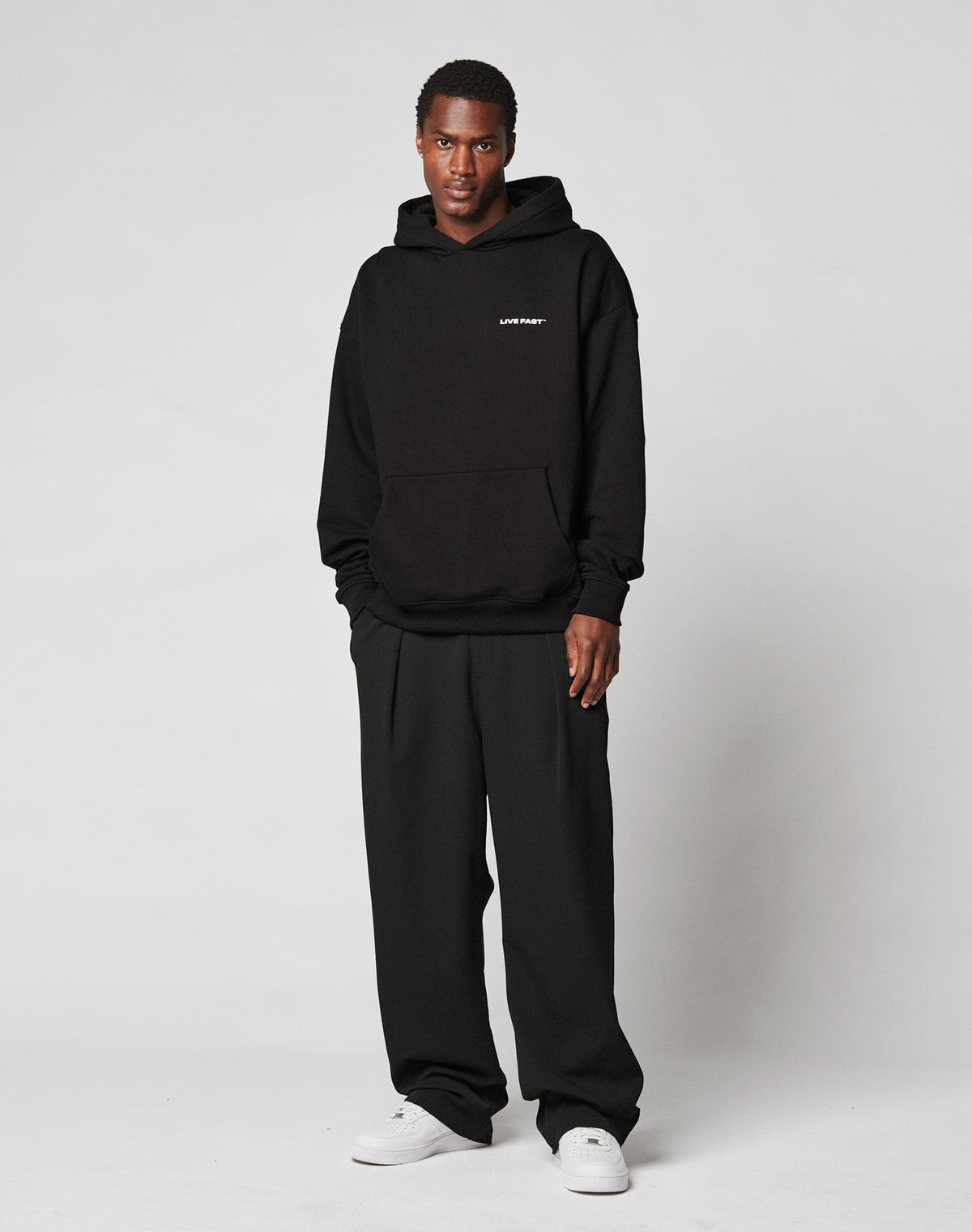 Against a plain white background, a person wears the LIVE FAST DIE YOUNG Live Fast Plain Hooded in oversize black with a small white logo, paired with matching wide-leg pants and white sneakers. The soft fabric is a Baumwolle-Polyester blend.