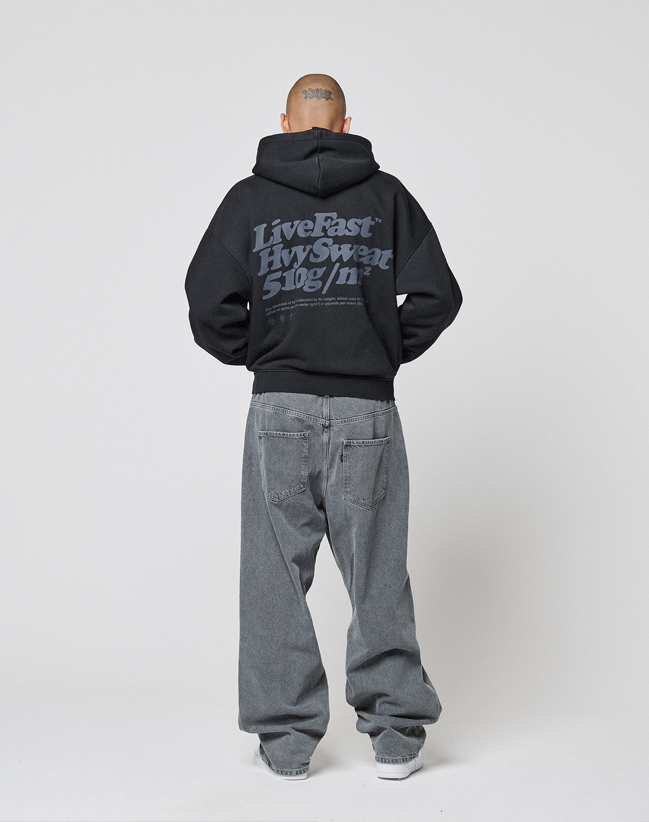 A person with a shaved head, facing away, wears an oversize fit black LIVE FAST DIE YOUNG Basic 510 Hooded Zip hoodie and loose gray vintage-wash jeans, standing against a plain white background.
