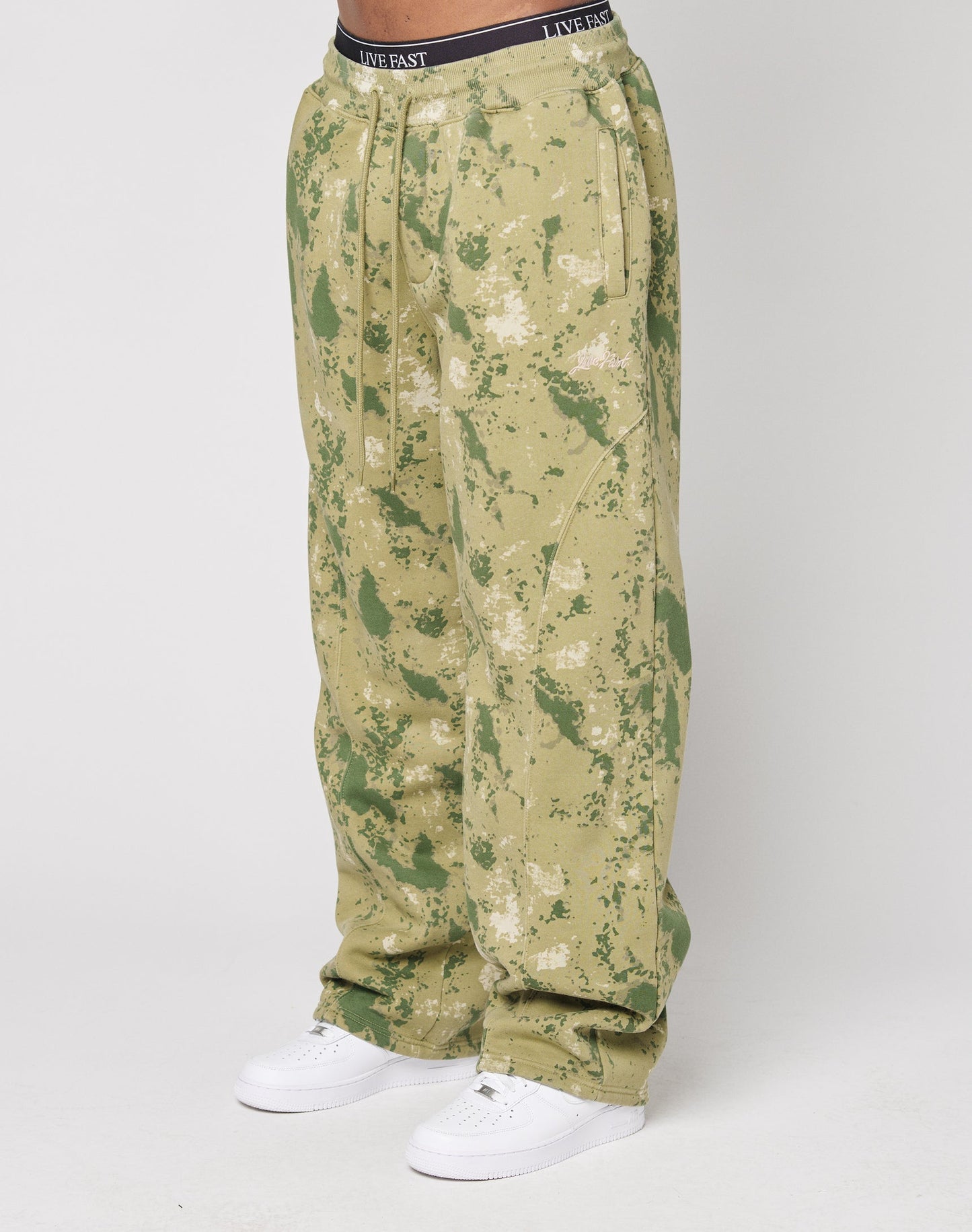 A person wears LIVE FAST DIE YOUNGs Camo Wide Jogger—loose-fitting, green and tan camouflage pants with a wide fit—paired with white sneakers and a visible black LIVE FAST waistband, set against a plain white background.