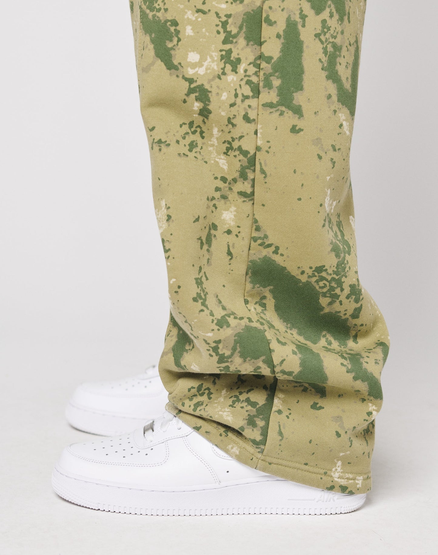 A person stands on a white surface wearing LIVE FAST DIE YOUNG’s Camo Wide Jogger, featuring olive green camo pattern, wide fit, and Elastikbund. The pants partly cover clean white sneakers; only the lower legs and feet are visible.