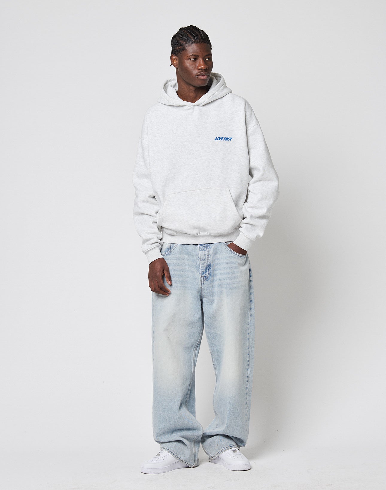 A man in a LIVE FAST DIE YOUNG LFDY Globe Hooded oversize light gray Baumwolle hoodie, loose light blue jeans, and white sneakers stands against a plain white background, gazing to the side with one hand in his pocket.