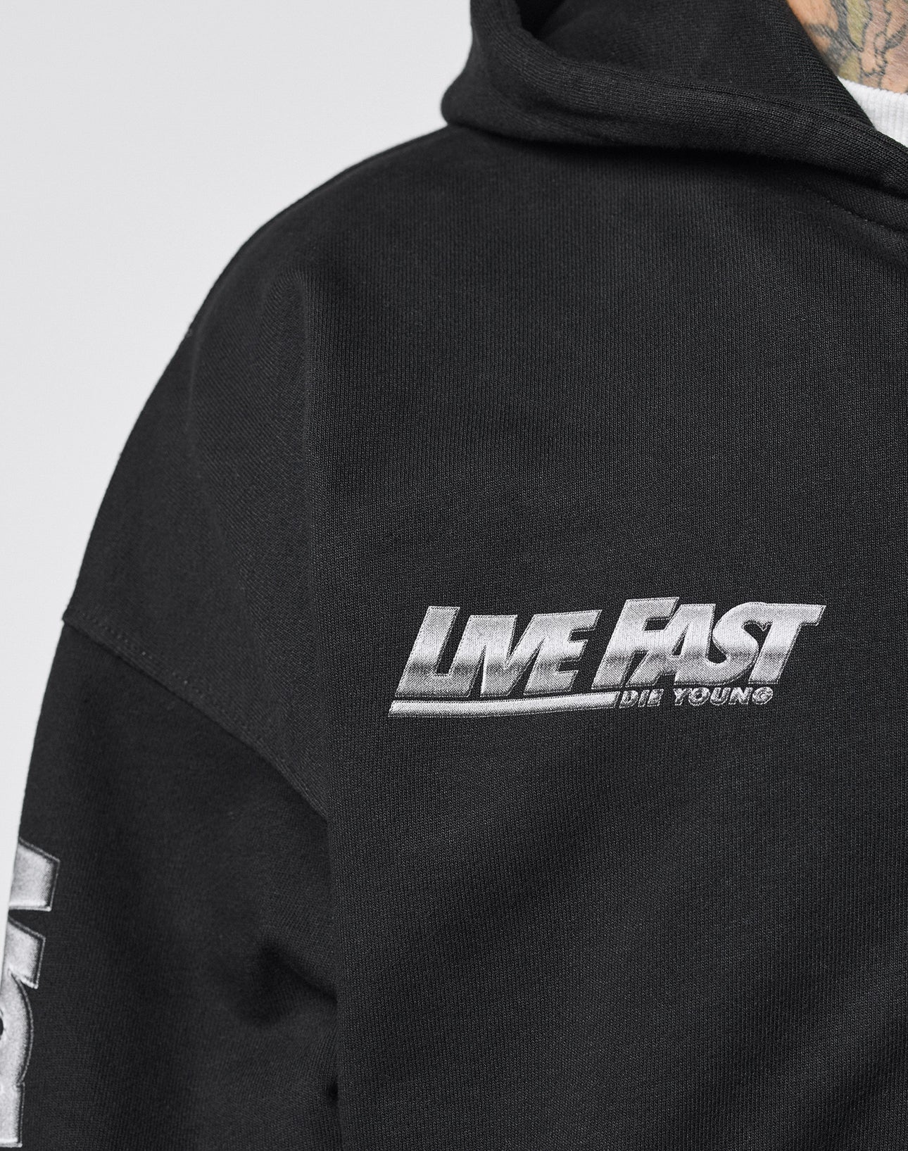 Close-up of the black LFDY Team Hooded Zip by LIVE FAST DIE YOUNG, featuring LIVE FAST DIE YOUNG embroidered in white on the chest, an oversize fit, and a glimpse of a tattooed neck and shoulder.