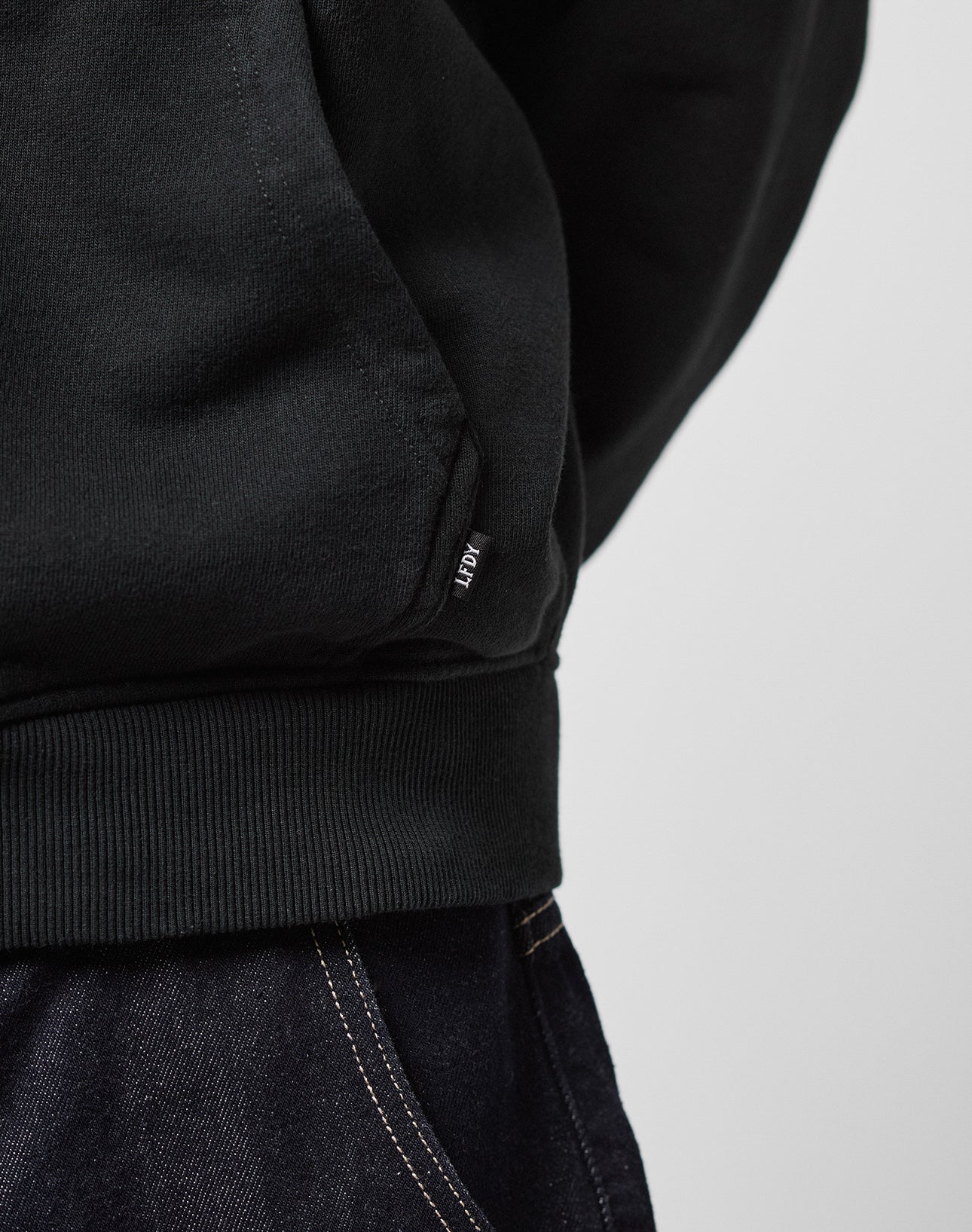 Close-up of a person wearing the LFDY Team Hooded Zip by LIVE FAST DIE YOUNG, an oversized black sweatshirt with ribbed hem, small ADER tag, visible Flaggen-Stick am Ärmel, paired with dark denim jeans featuring white stitching.