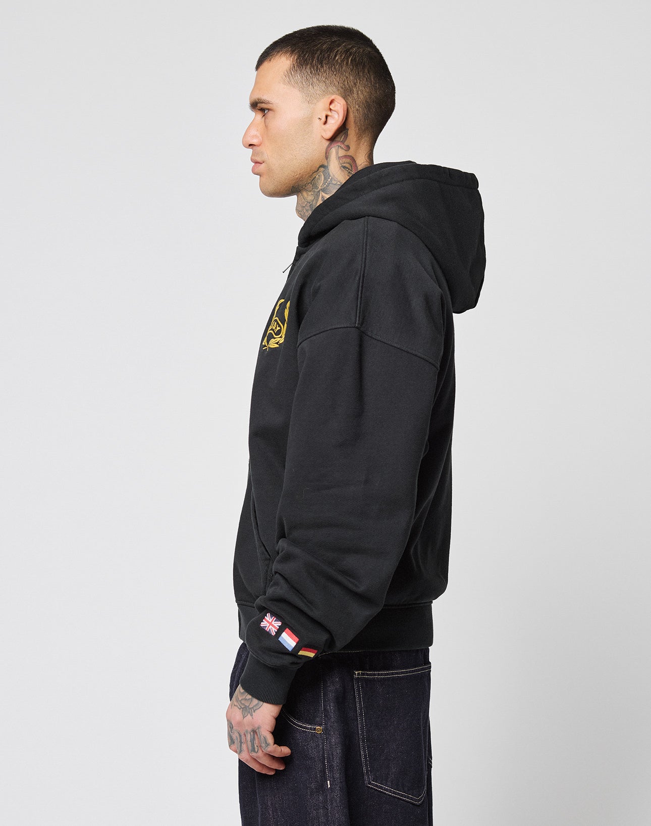 A person with short hair and visible tattoos wears the LIVE FAST DIE YOUNG LFDY Team Hooded Zip in Oversize Fit and dark jeans, standing in profile against a plain light gray background. The hoodie features a flag embroidery on the sleeve.