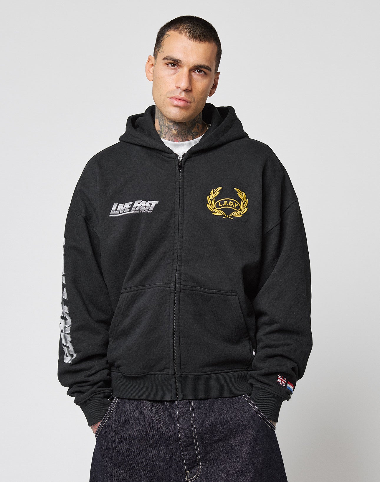 A man stands facing forward with hands in pockets, wearing the black LFDY Team Hooded Zip by LIVE FAST DIE YOUNG featuring white and gold Multilogo Stickereien, against a plain light background.