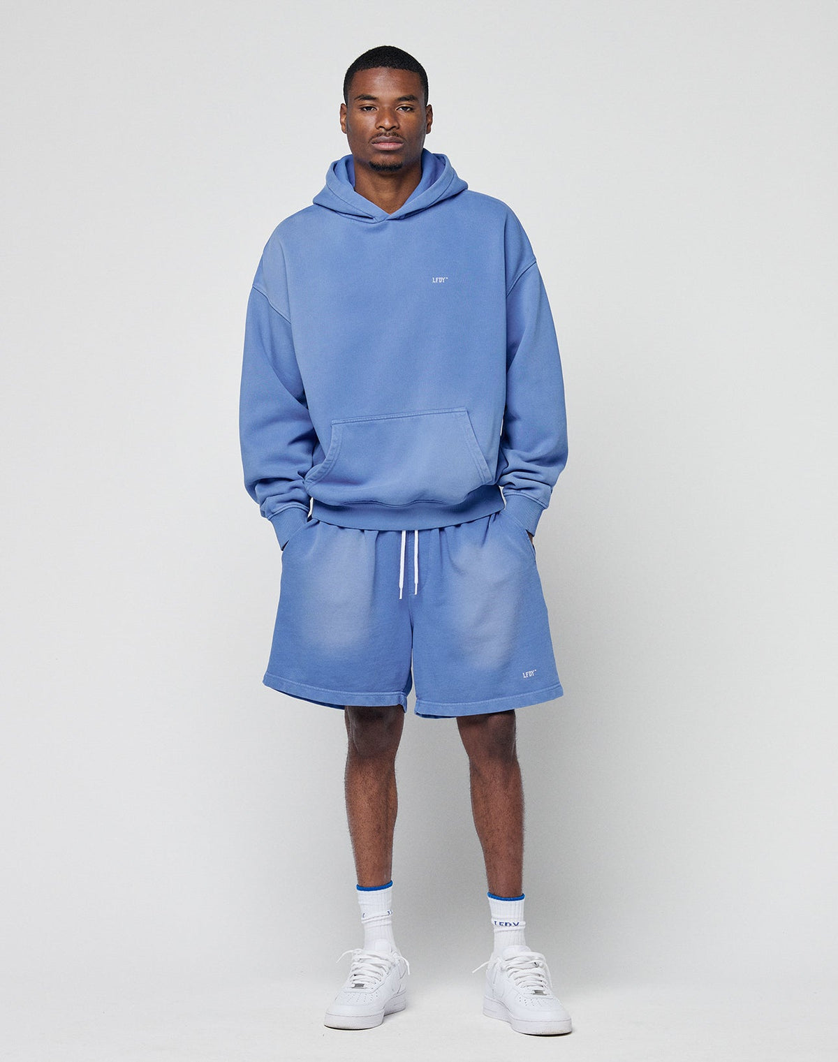 A man stands against a plain background wearing the LIVE FAST DIE YOUNG Basic Evidence Hooded light blue oversized hoodie and shorts set with a subtle faded look, paired with white crew socks and sneakers, looking at the camera with hands in his pockets.