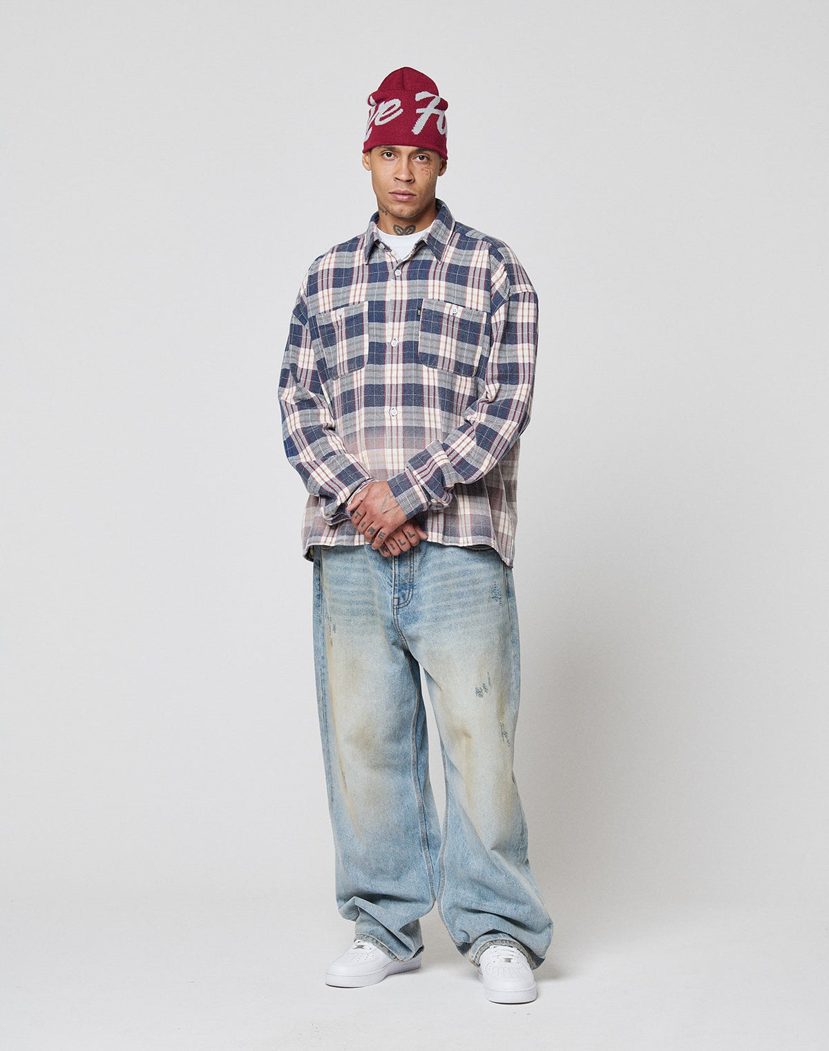 A person stands against a plain background wearing a maroon beanie, loose-fitting faded jeans, white sneakers, and the LIVE FAST DIE YOUNG Vintage Flannel shirt with hands clasped and a neutral expression.