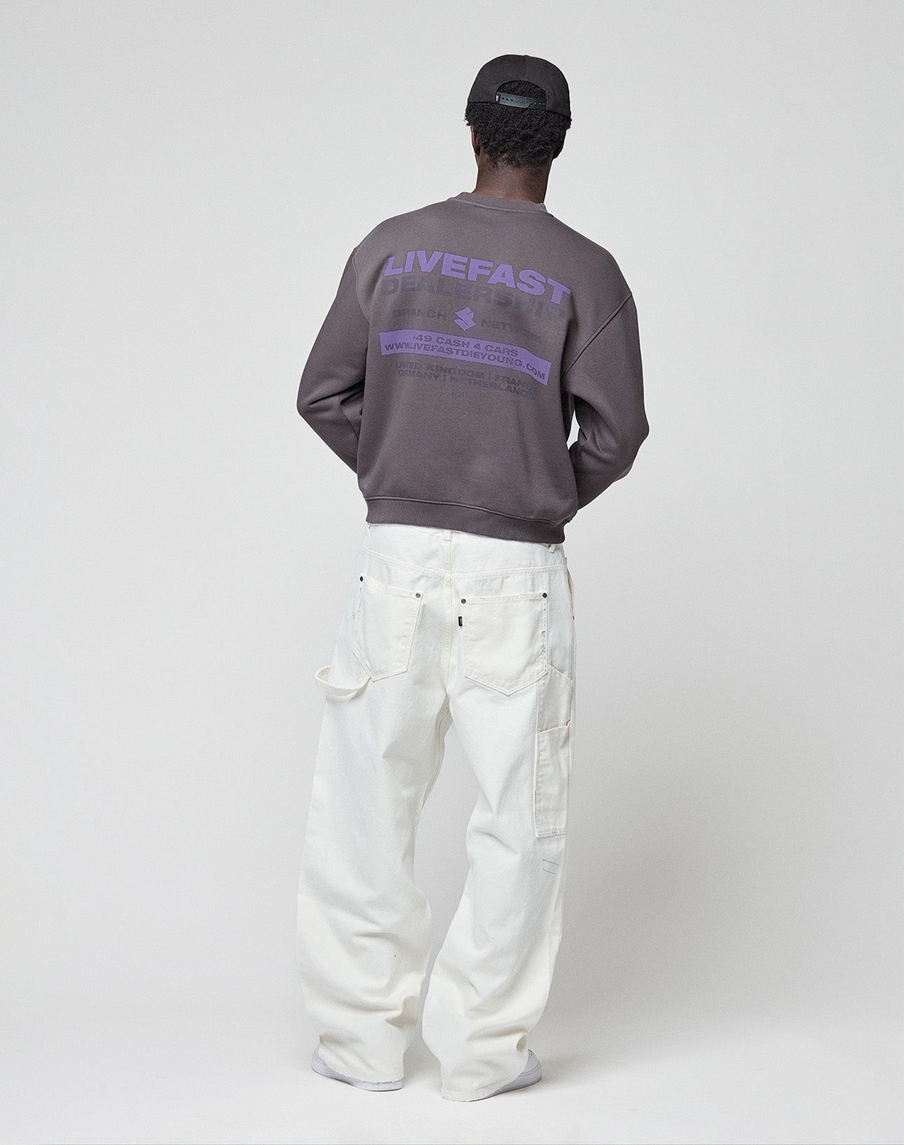 A person stands facing away in a dark cap, white loose pants, and the LIVE FAST DIE YOUNG "LFDY Network Sweater," an oversized purple cotton sweatshirt with LIVEFAST front and back print, against a plain light studio background.