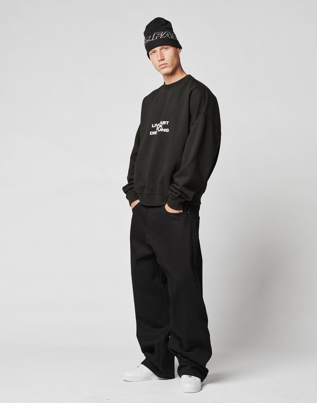 A person wearing a LIVE FAST DIE YOUNG Fuck Die Young Sweater, black beanie, loose black pants, and white sneakers stands against a plain white background with hands in pockets.