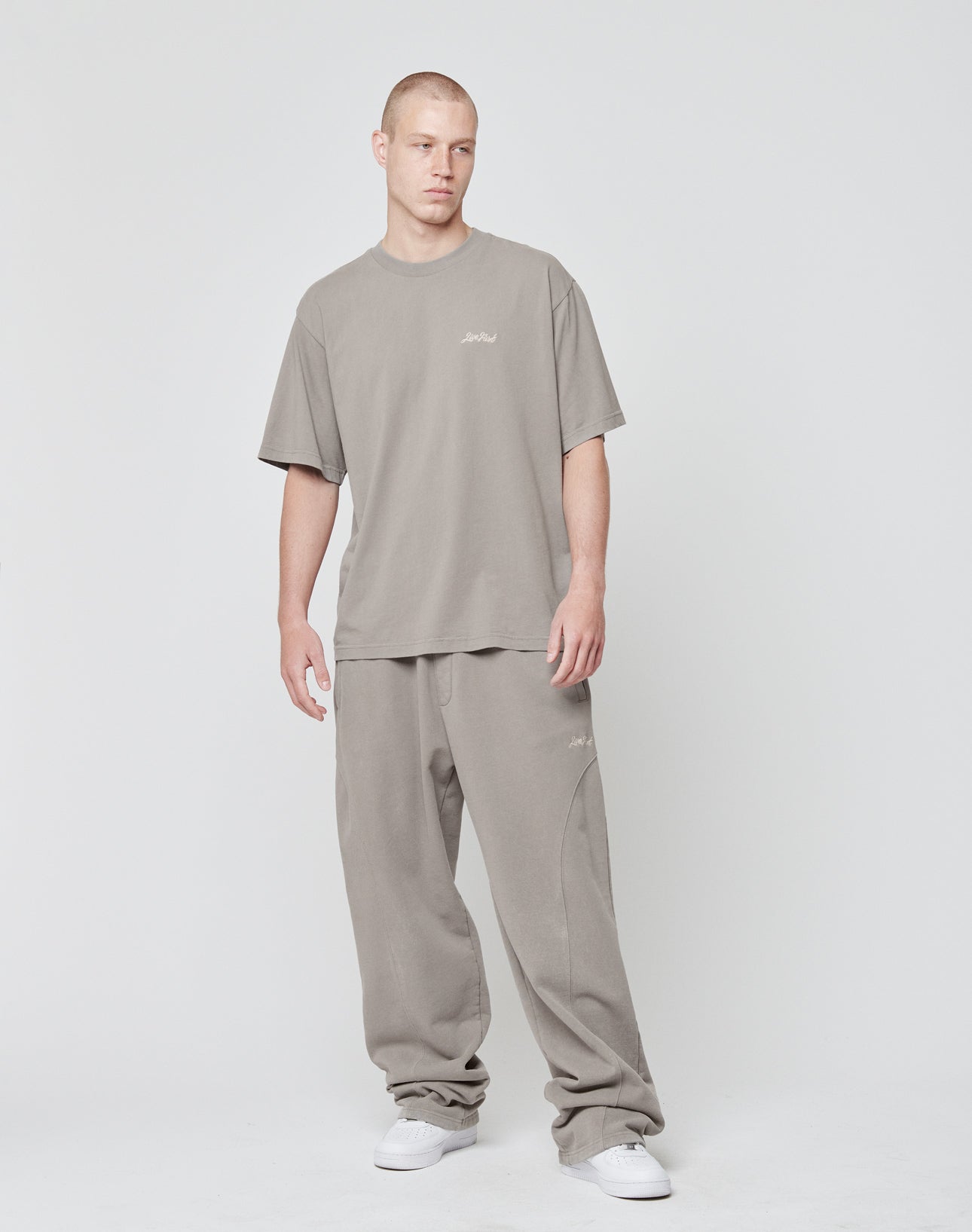 A man with a neutral expression stands against a white background, wearing the LIVE FAST DIE YOUNG Basic Script Tee in oversized taupe Vintage-Waschung and matching baggy sweatpants, paired with white sneakers. His arms rest relaxed at his sides.
