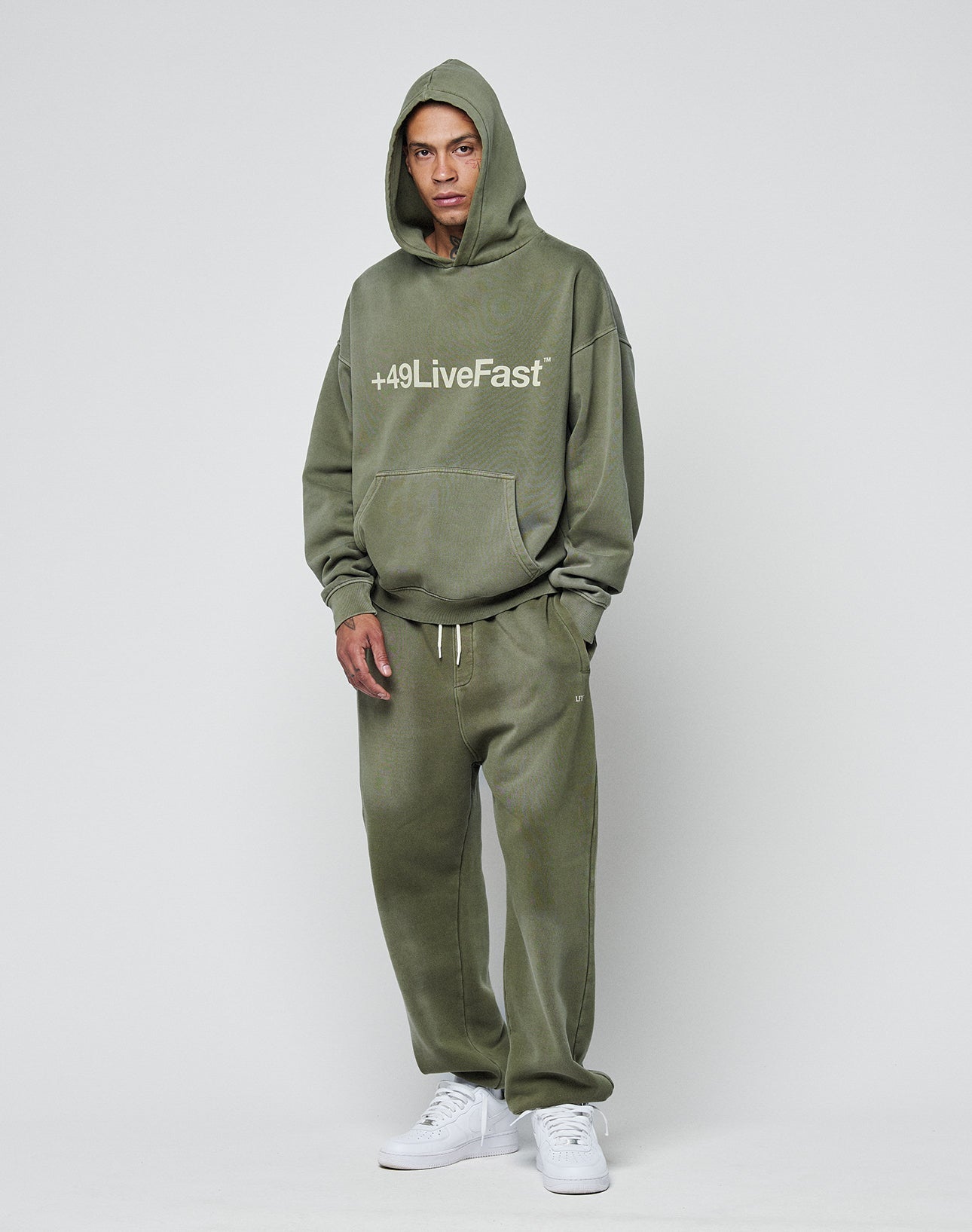 A person wears the LIVE FAST DIE YOUNG Basic +49 Hooded in olive green with matching Oversize Fit sweatpants and white sneakers, standing against a plain light gray background.