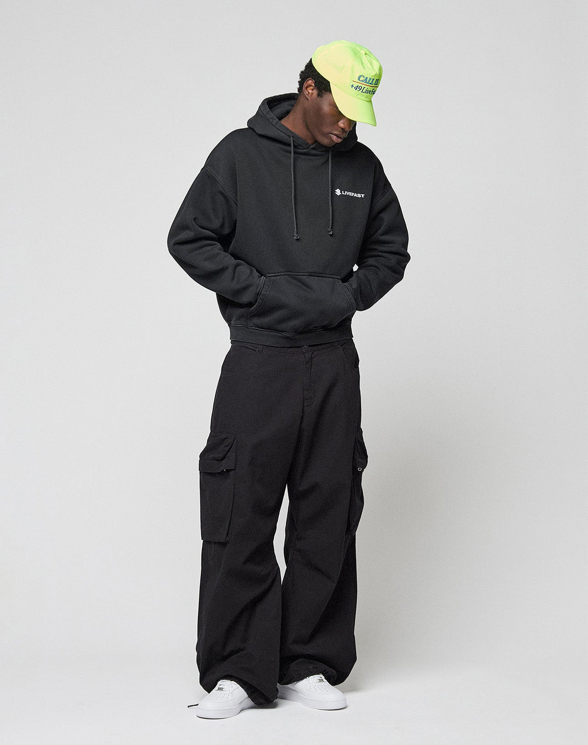 A person in a black LIVE FAST DIE YOUNG LFDY Dealership Hooded with front and back print, black cargo pants, white sneakers, and a neon green cap stands against a light background, hands in the hoodie pocket and gazing slightly down.