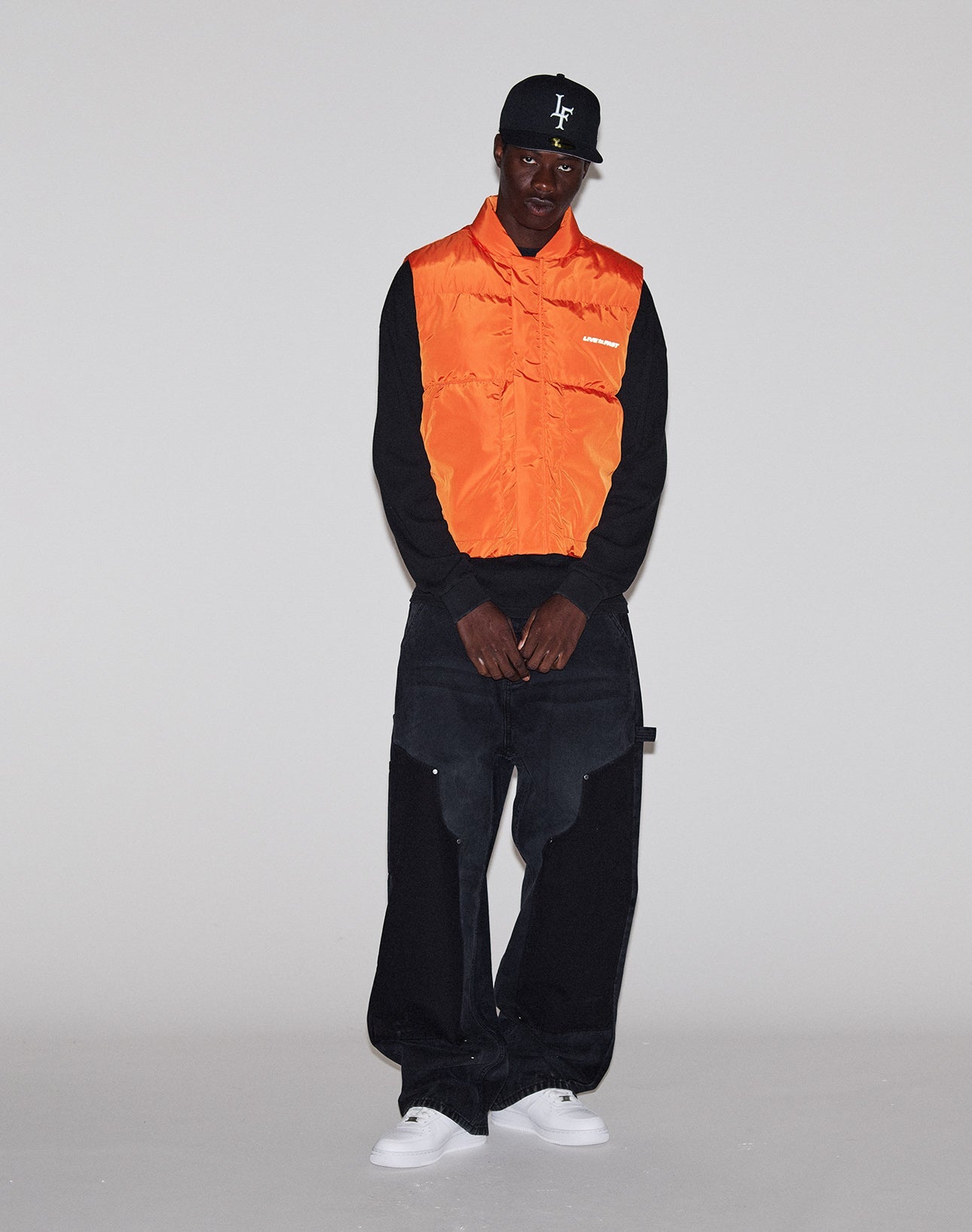 A man stands against a plain background, wearing the LIVE FAST DIE YOUNG Live Fast Cropped Vest in orange over a black long-sleeve shirt, with a black cap, oversized black pants, and white sneakers.
