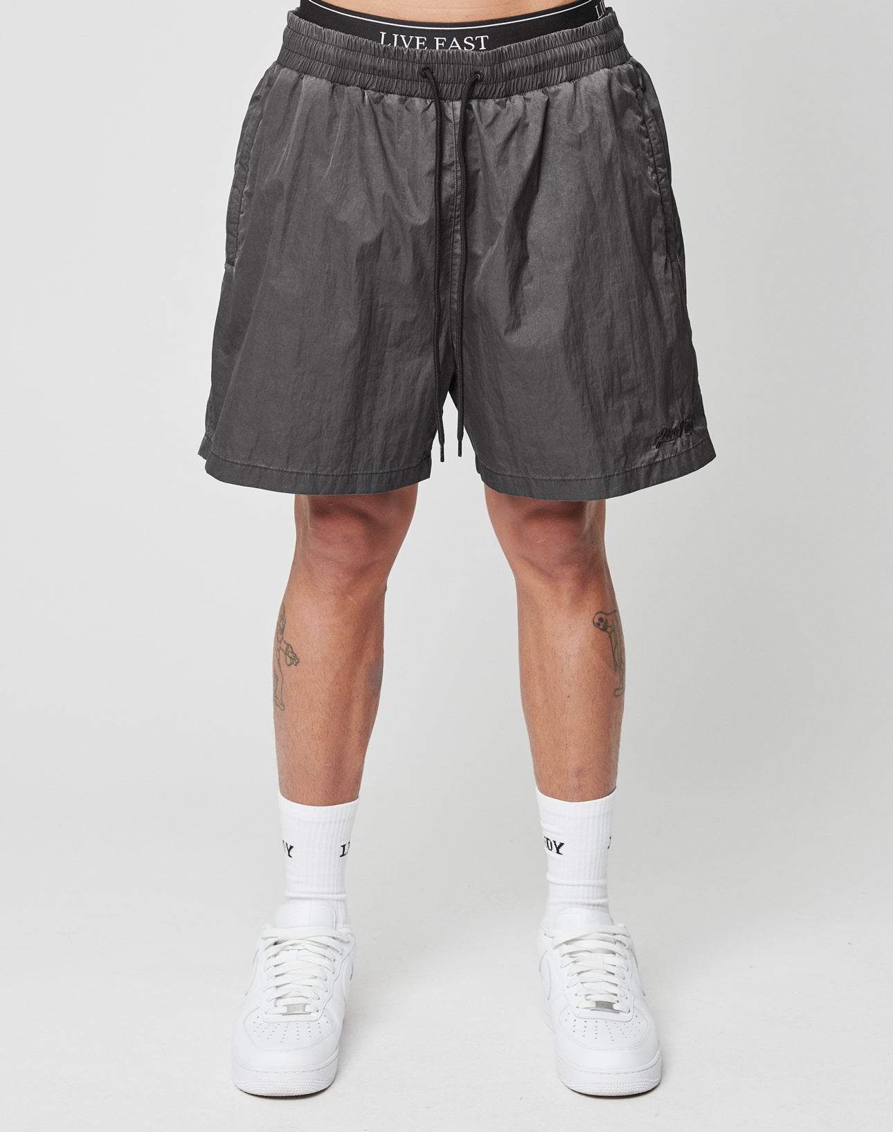 A person stands against a plain background wearing LIVE FAST DIE YOUNG Live Fast Swimshorts with a logo waistband. The tinted black polyamide shorts are regular fit. Only their lower body is visible, along with white socks and sneakers.