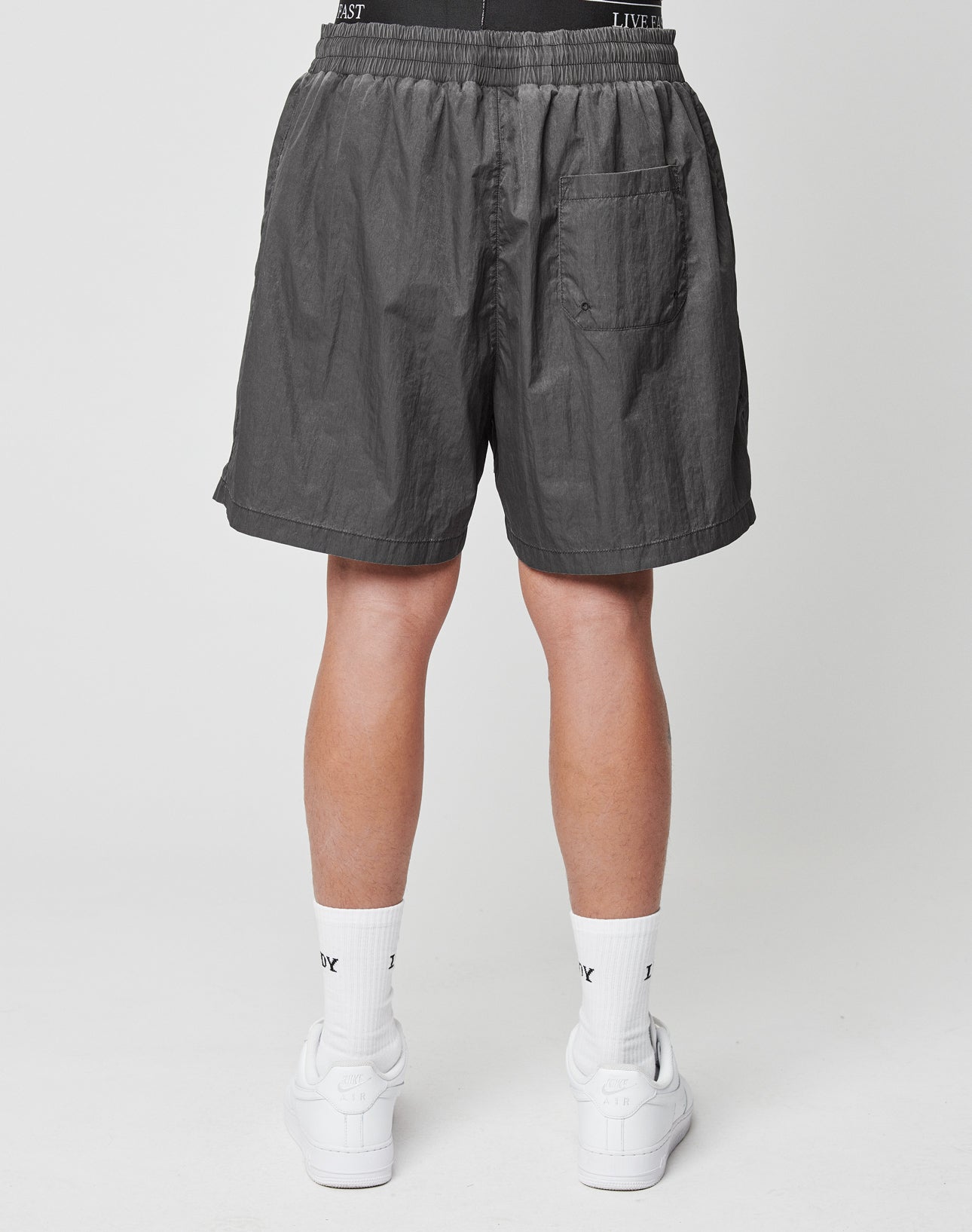 A person is seen from behind wearing LIVE FAST DIE YOUNG Live Fast Swimshorts in Tinted Black, featuring a regular fit, elastic waistband, and single back pocket, paired with white crew socks and sneakers on a plain white background.