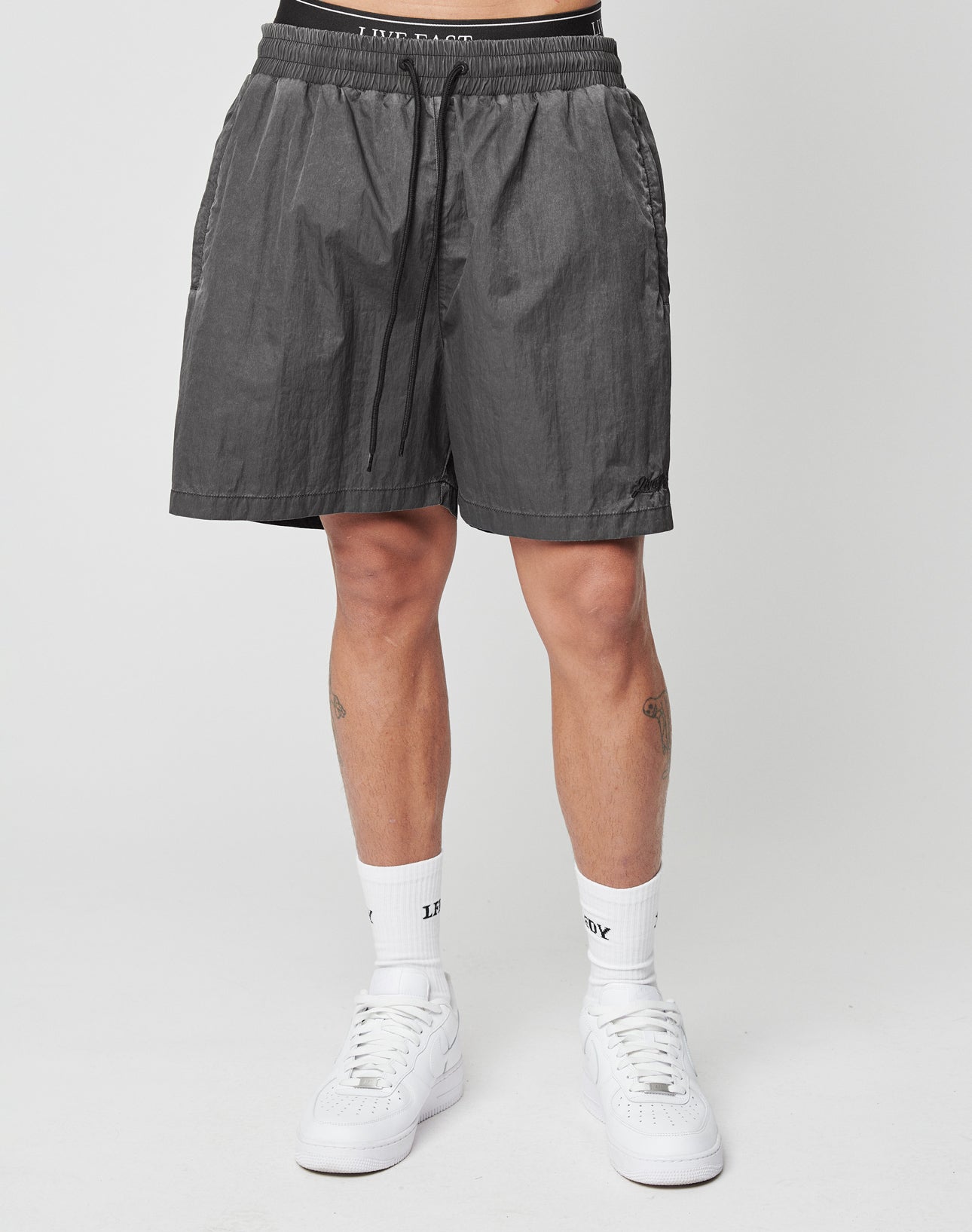 A person wearing LIVE FAST DIE YOUNG Live Fast Swimshorts in black, paired with white crew socks and white sneakers, stands against a plain light background. Only the lower half of the body is visible.