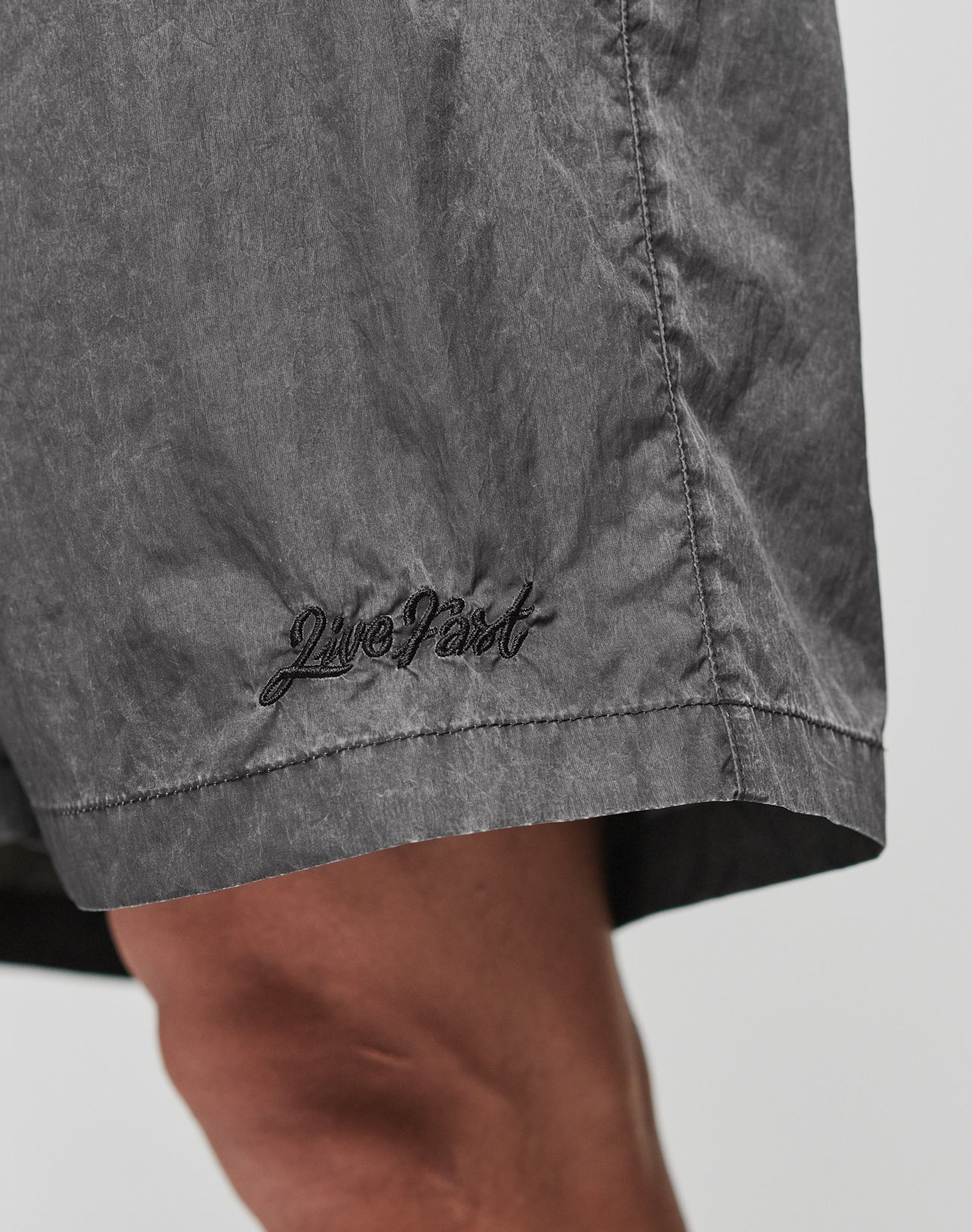 Close-up of a person wearing LIVE FAST DIE YOUNG Live Fast Swimshorts in gray, Regular Fit, with Live Fast embroidered in black near the hem. The background is plain and light-colored.