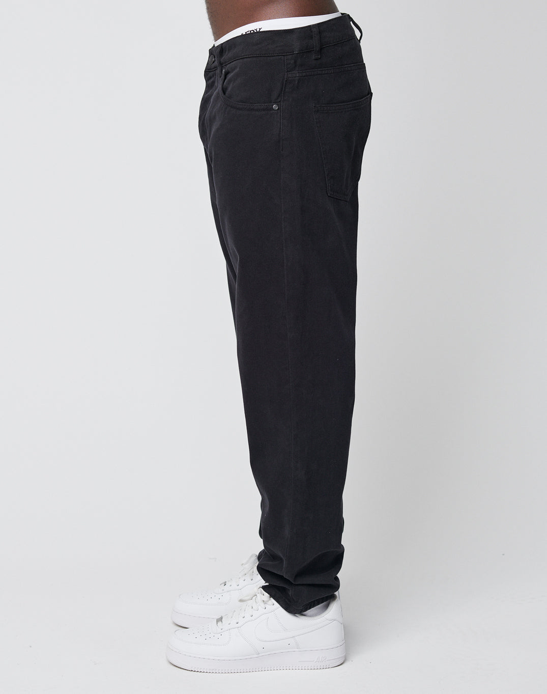 A person’s lower half is shown in profile, wearing LIVE FAST DIE YOUNG Regular Denim—black, regular fit with a tapered leg—and white sneakers, set against a plain white background.