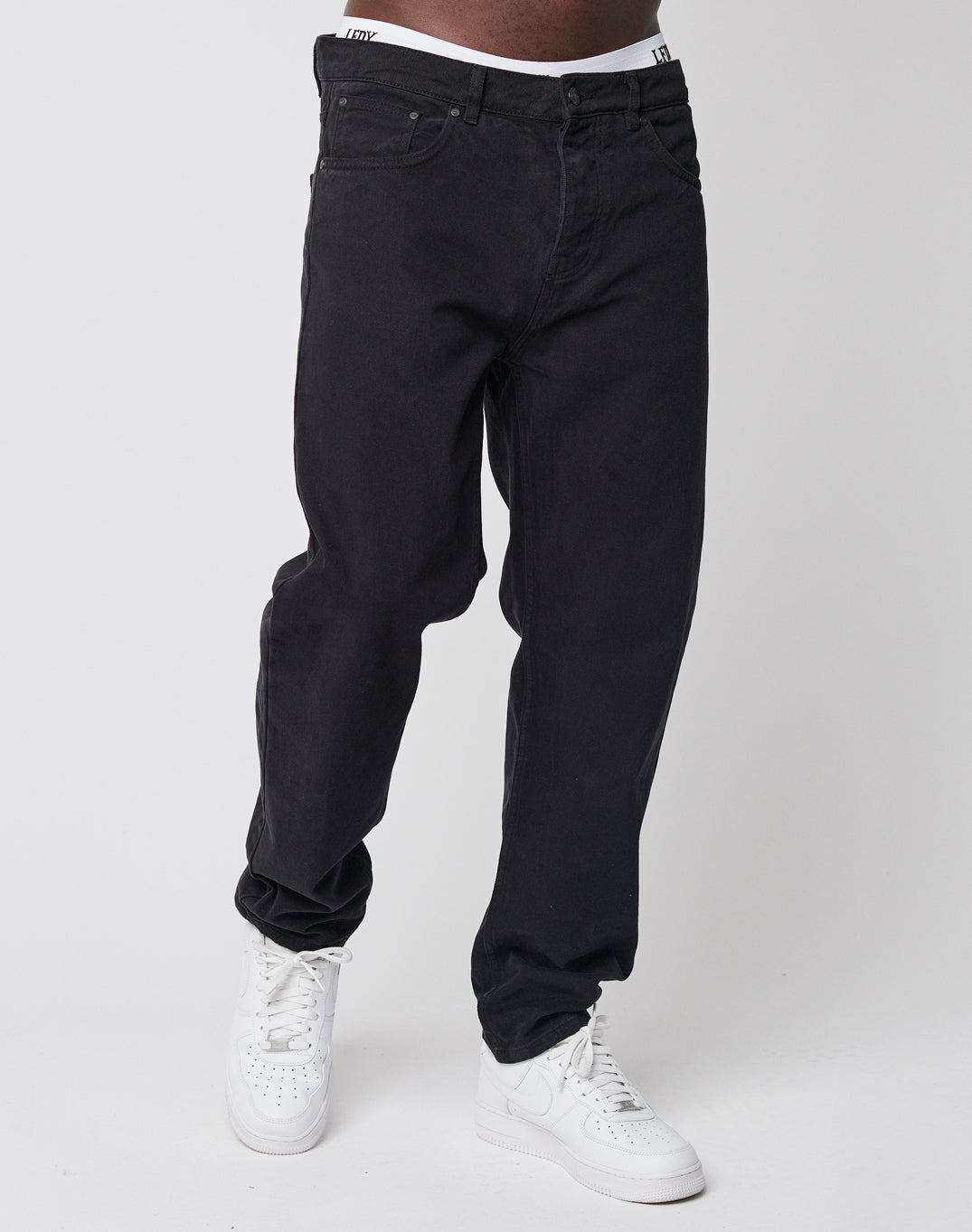 A person wears LIVE FAST DIE YOUNG Regular Denim—loose, tapered black jeans paired with white boxer briefs and sneakers. The mid-chest-down photo is set against a plain white background.