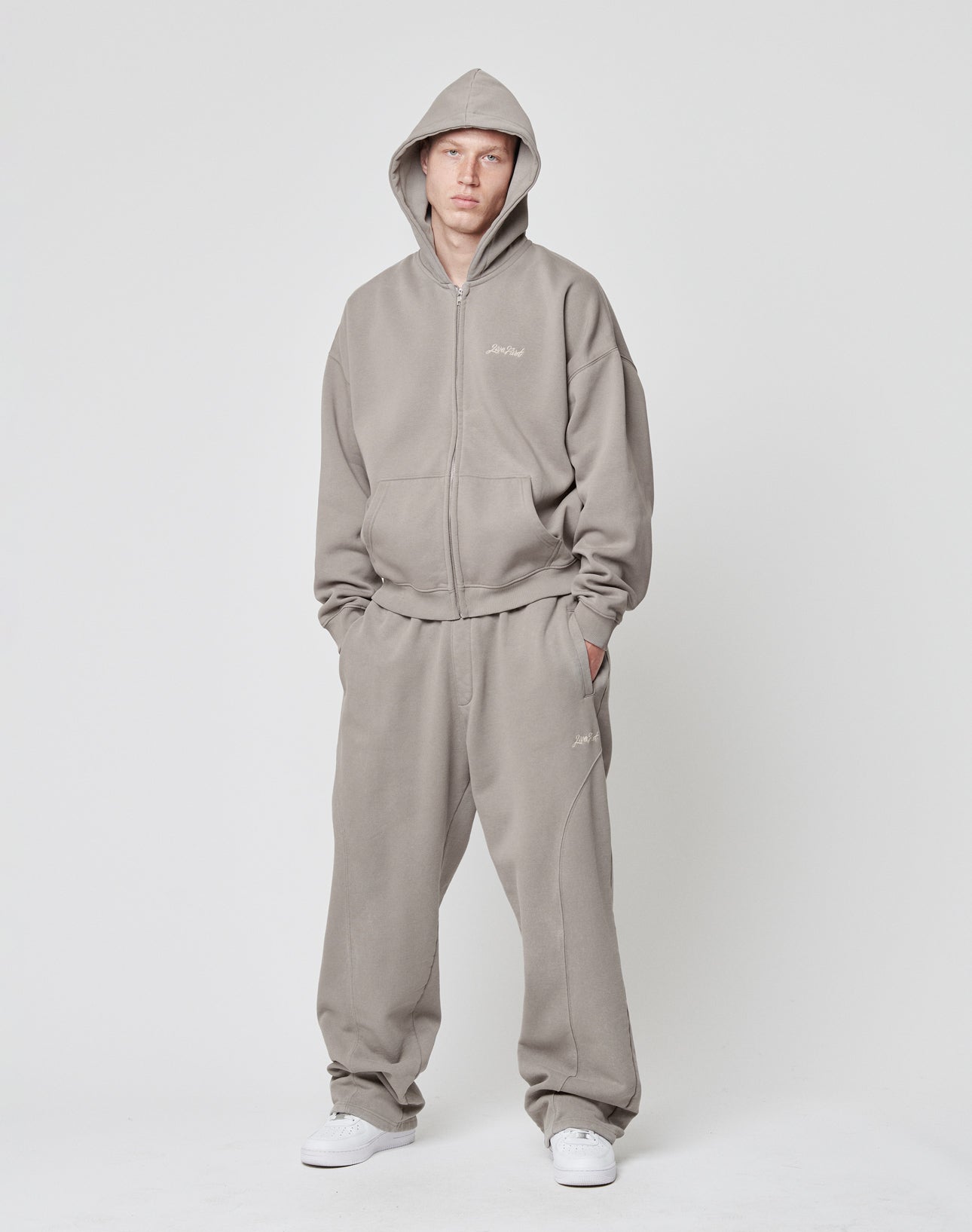 Eine Person trägt den Basic Script Hooded Zip von LIVE FAST DIE YOUNG in Hellgrau sowie passende, weite Sweatpants. Mit aufgesetzter Kapuze und Händen in den Taschen posiert sie vor weißem Hintergrund; weiße Sneakers runden das Outfit ab.