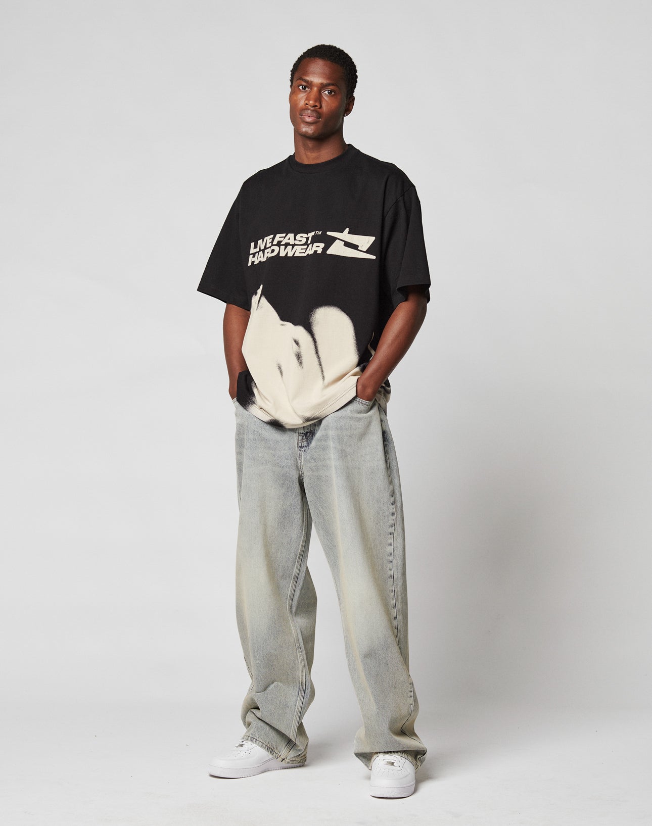 Against a plain light gray background, a person wears the LFDY Mystic Tee by LIVE FAST DIE YOUNG in Oversize Fit with front and back print, baggy light-wash jeans, and white sneakers, hands in pockets.