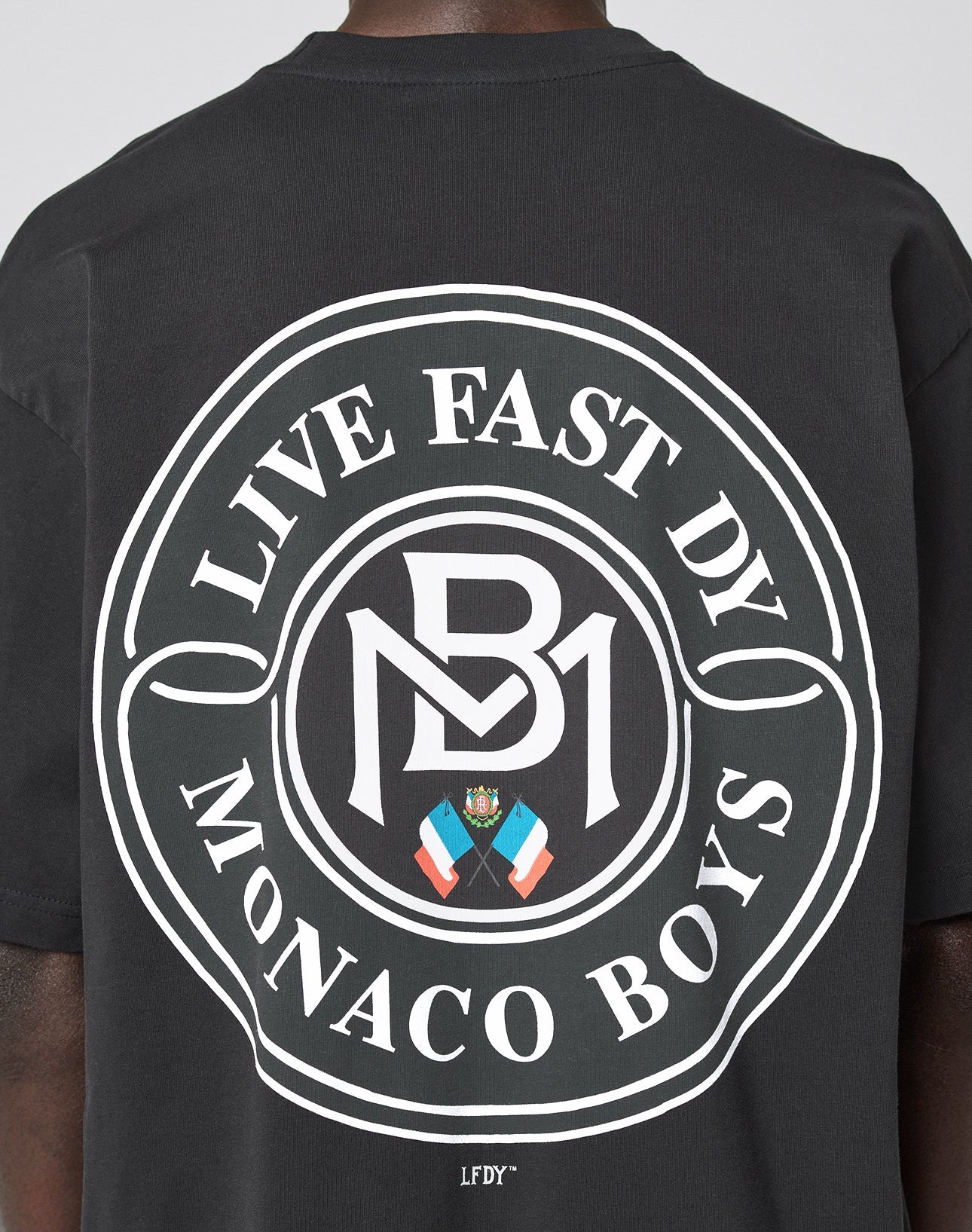 Someone wearing the LFDY Monaco Boys Tee: an oversized black shirt with a large circular white graphic on the back reading LIVE FAST DY MONACO BOYS, featuring a crest, flags, and MB initials. Vintage effect, 100% cotton.