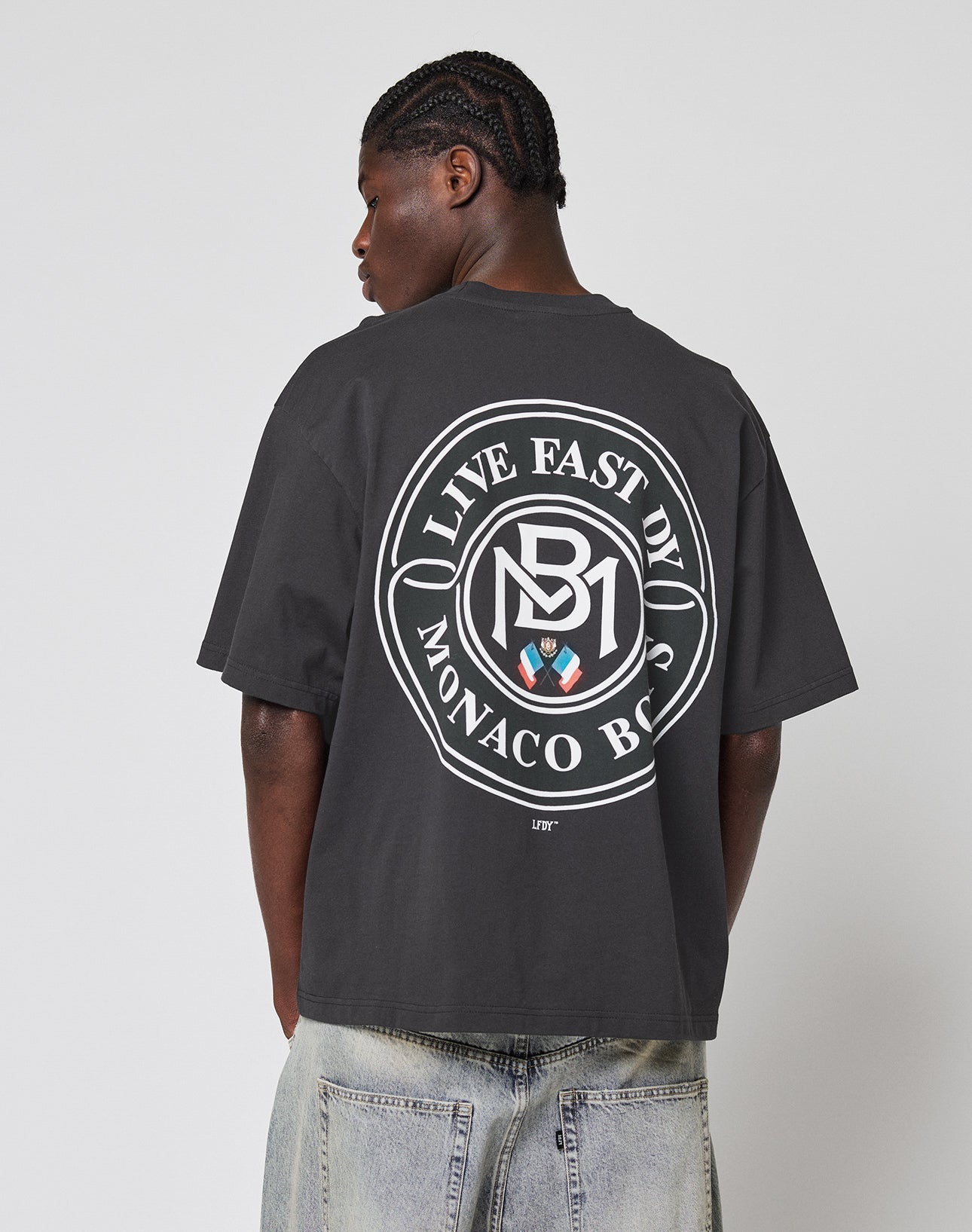 A person with braided hair and light denim jeans faces away, wearing the LFDY Monaco Boys Tee by LIVE FAST DIE YOUNG in oversized fit, vintage optic, featuring a large circular back graphic with LIVE FAST, DIE YOUNG MONACO BOYS text.
