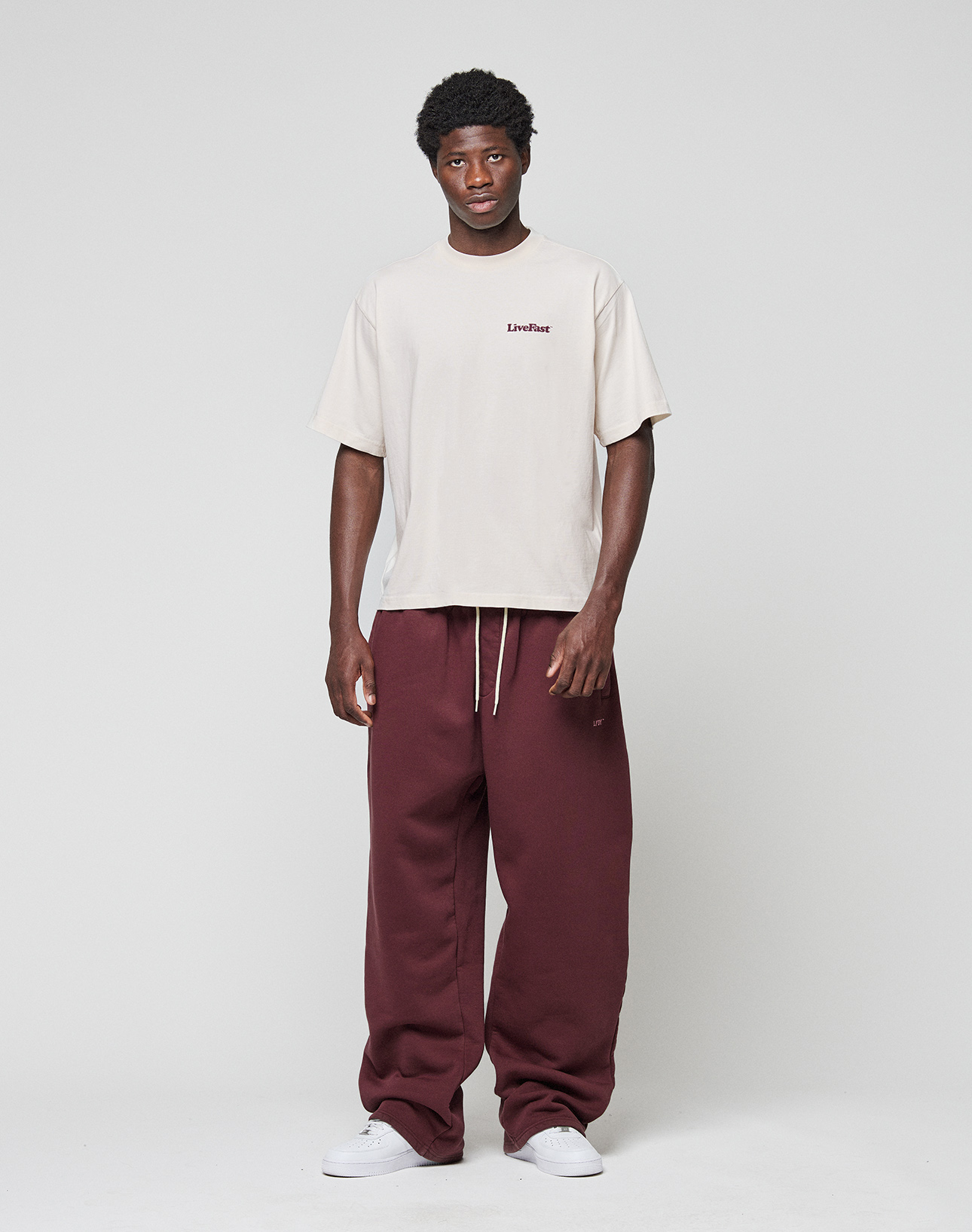 A person stands against a plain background wearing the LIVE FAST DIE YOUNG Basic 210 Tee in oversize beige with front and back Livefast print, paired with loose maroon drawstring sweatpants and white sneakers.