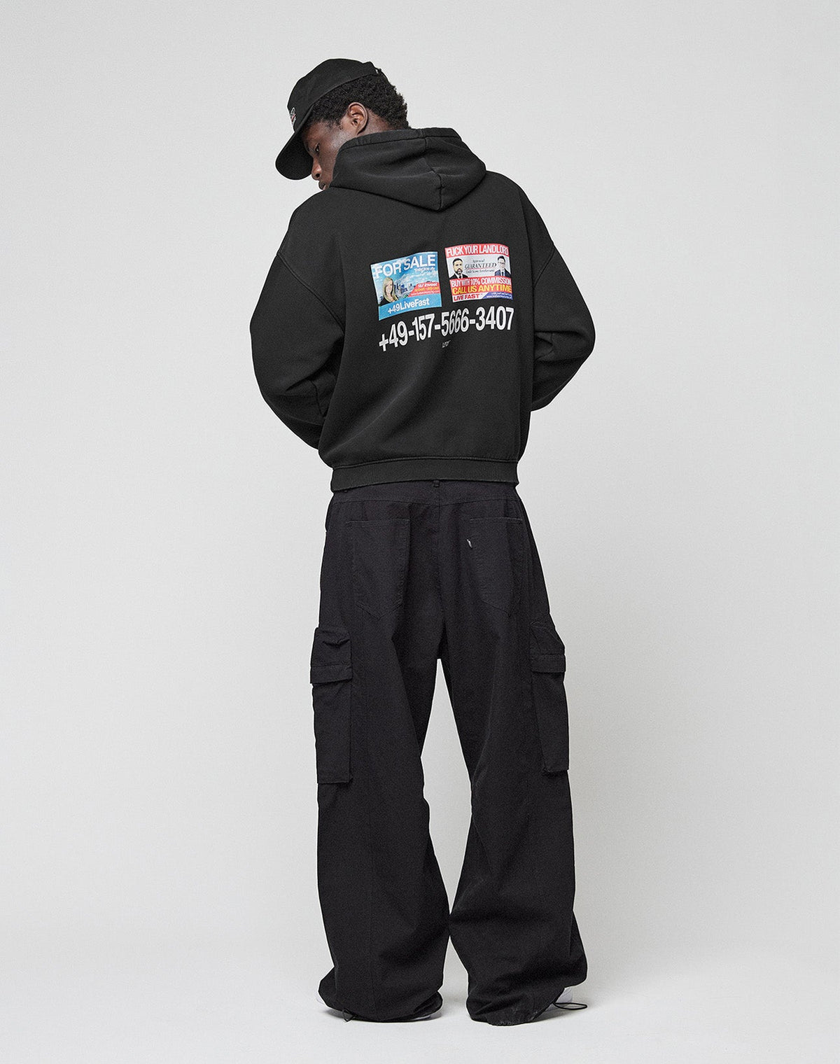 A person wearing the LIVE FAST DIE YOUNG Live Fast Call Us Hooded, an oversize black cotton hoodie with colorful prints, black cargo pants, and a cap stands facing away against a plain light background.