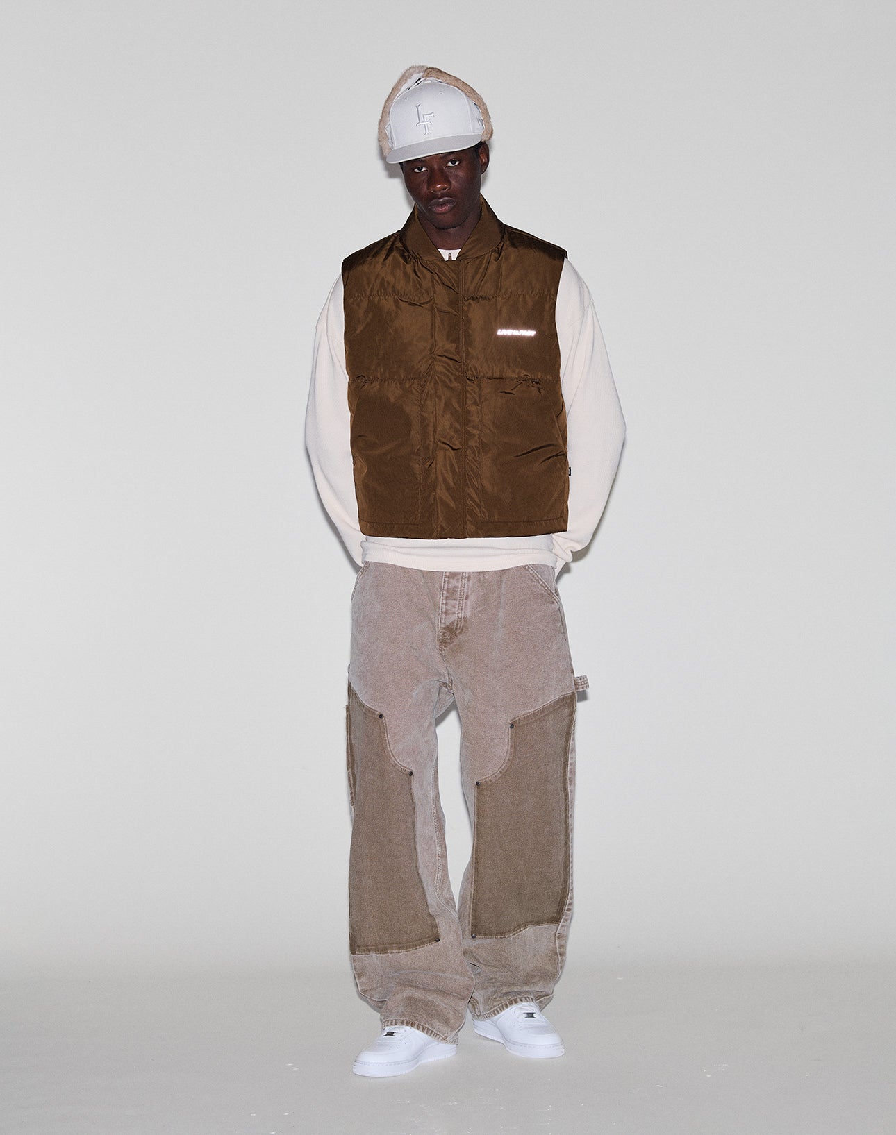 A person stands against a plain light background wearing the LIVE FAST DIE YOUNG Live Fast Cropped Vest in brown over a cream long-sleeve shirt, beige patchwork pants, a beige hat, and white sneakers.
