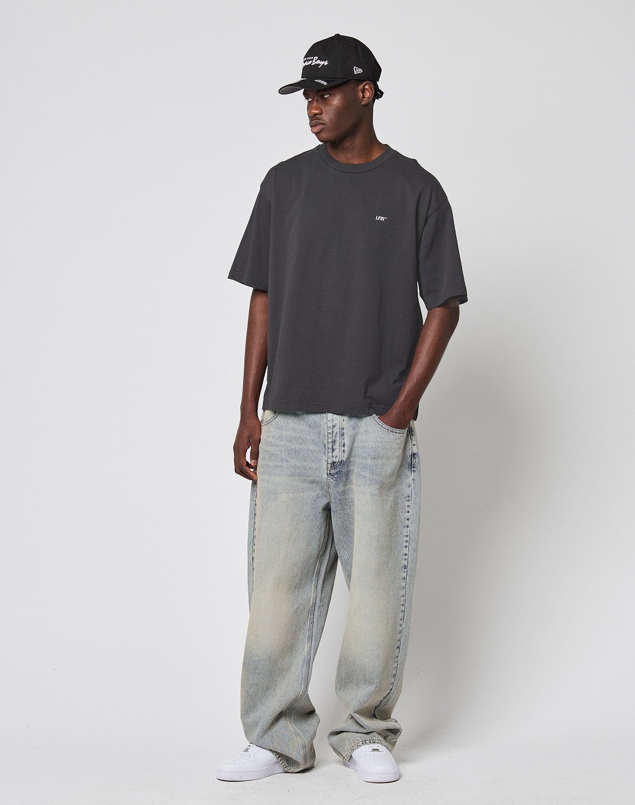 A person in a black cap, LIVE FAST DIE YOUNG LFDY Monaco Boys Tee in dark gray Oversize Fit, baggy light-wash jeans in Vintage Optik, and white sneakers stands against a plain white background with hands in pockets, looking to the side.