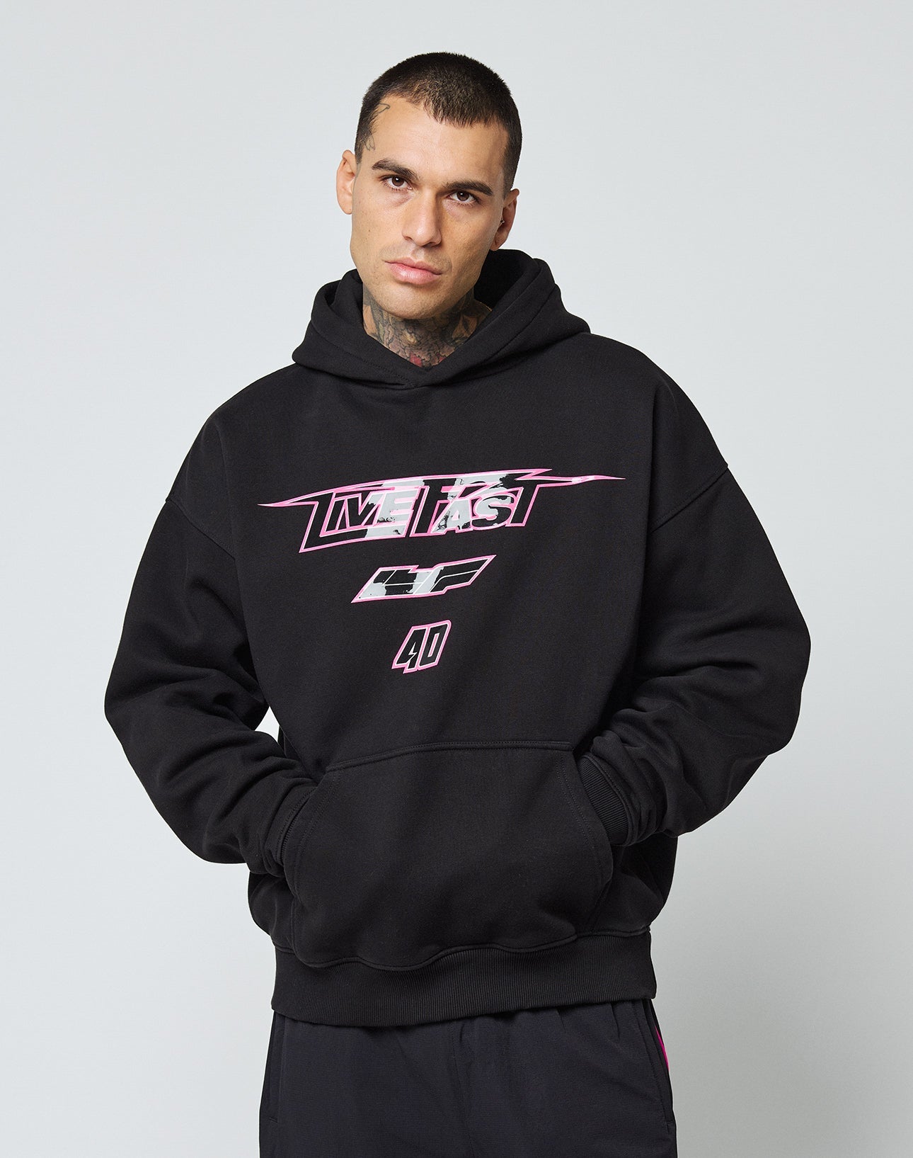 A person with short dark hair and neck and hand tattoos wears the LIVE FAST DIE YOUNG LFDY Triple Racing Hooded, an oversize black Baumwolle sweatshirt with multilogo pink and white graphic text and numbers on the front, standing against a plain light background.