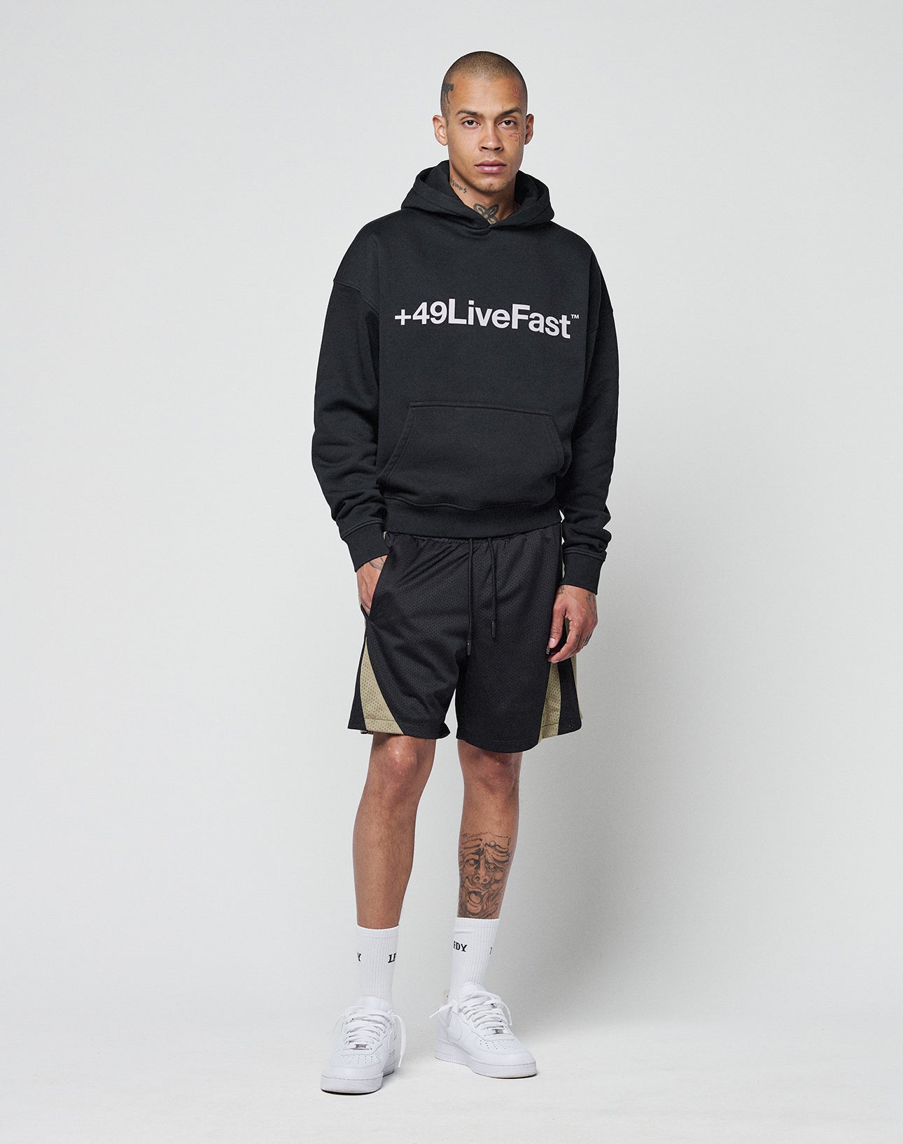 A man wears the LIVE FAST DIE YOUNG Basic +49 Hooded in black, featuring a +49LiveFast front print and oversize fit (Baumwolle-Polyester blend), paired with black shorts with beige side panels, white socks, and white sneakers.