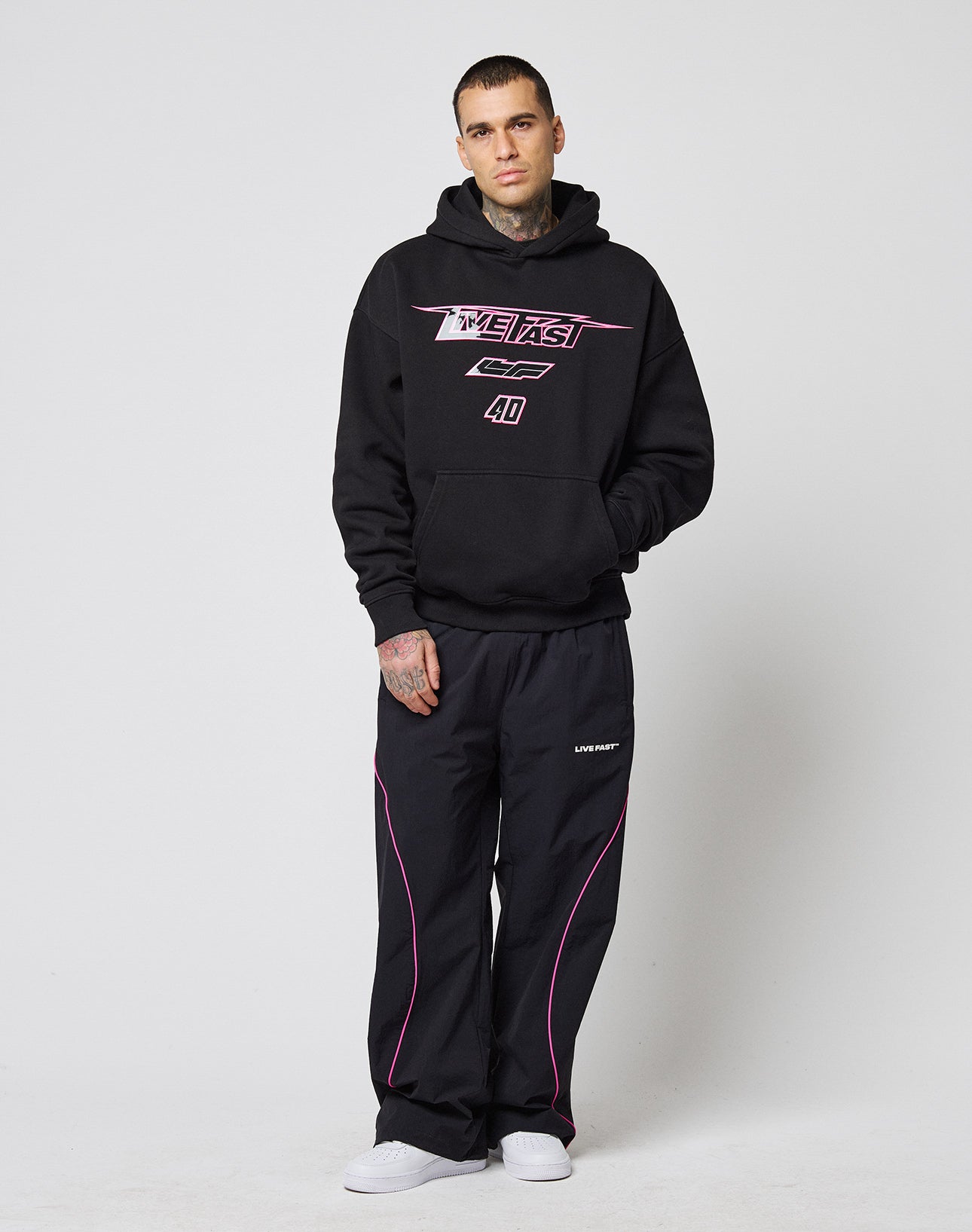 A person with short hair, tattoos, and a neutral expression wears an LFDY Triple Racing Hooded by LIVE FAST DIE YOUNG—an oversize black hoodie with pink graphics and multilogo details—paired with black pants with pink piping and white sneakers.