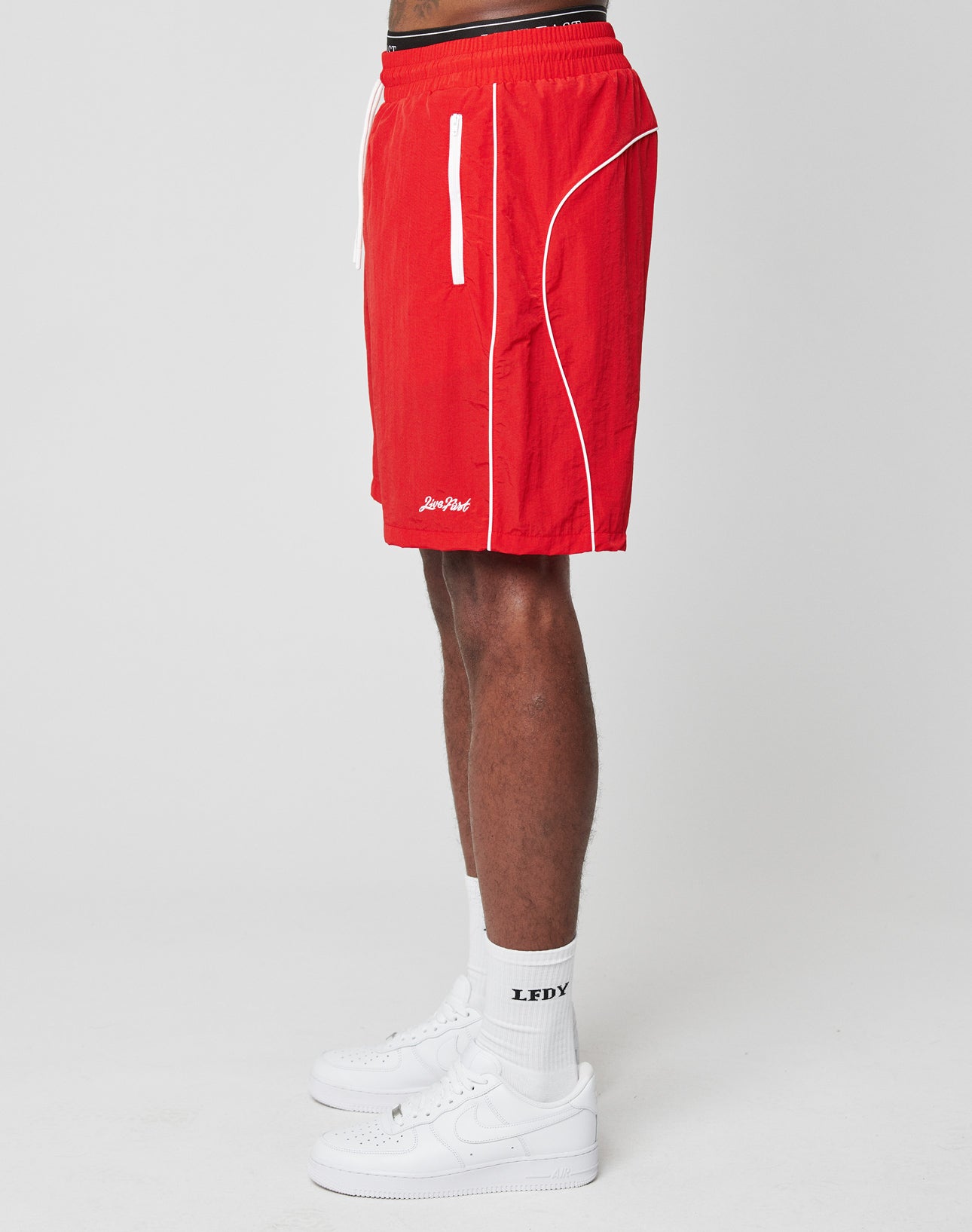 A person in bright red Beach Shorts by LIVE FAST DIE YOUNG, featuring an elastic waistband and white trim, along with LFDY white socks and white sneakers, stands in side profile against a plain white background.