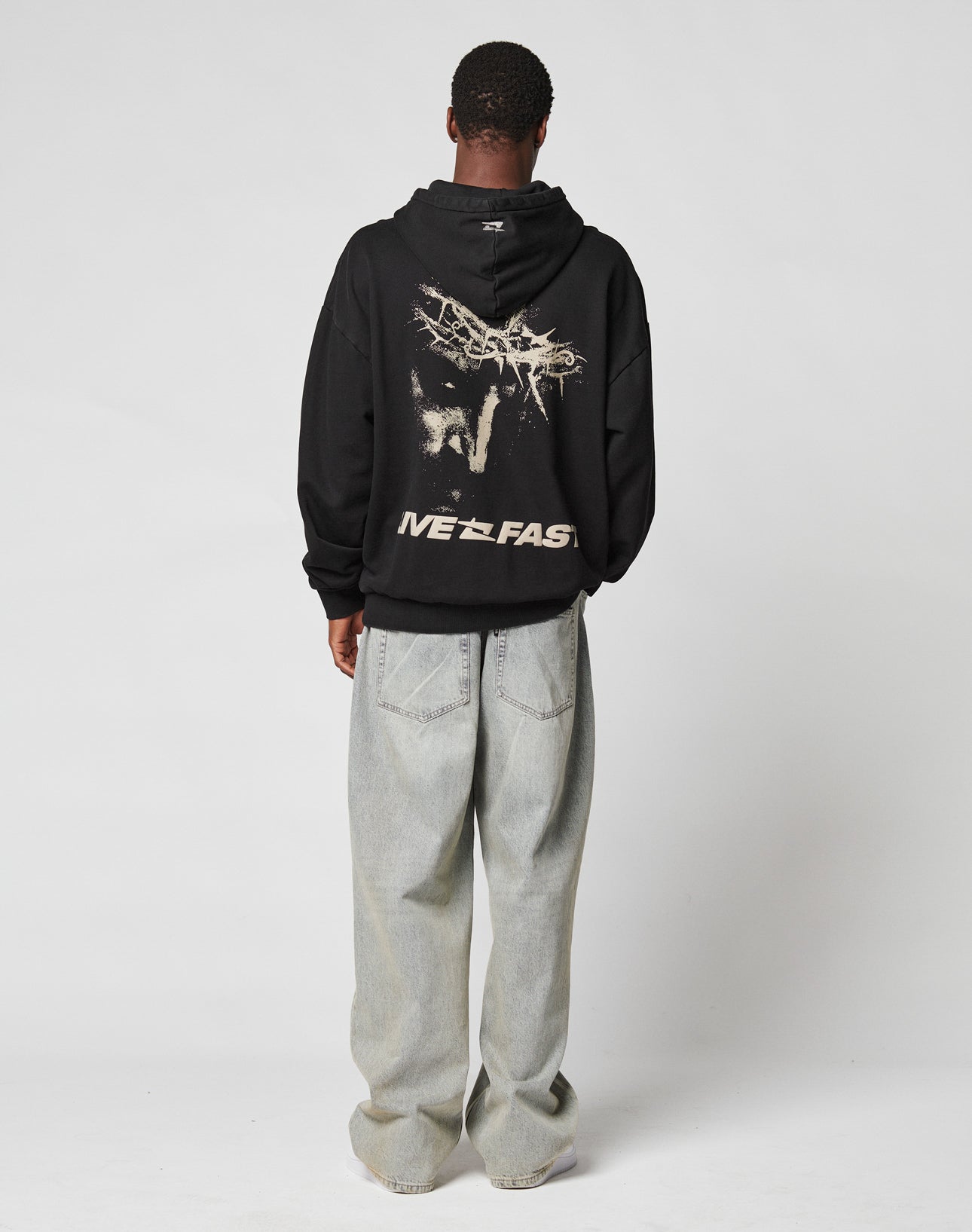 A person faces away wearing the LFDY Savior Hooded by LIVE FAST DIE YOUNG, an oversized black hoodie made from 100% Baumwolle with a tree, animal graphic, logo stick, and LIVE FAST on the back, paired with baggy light jeans and white sneakers.