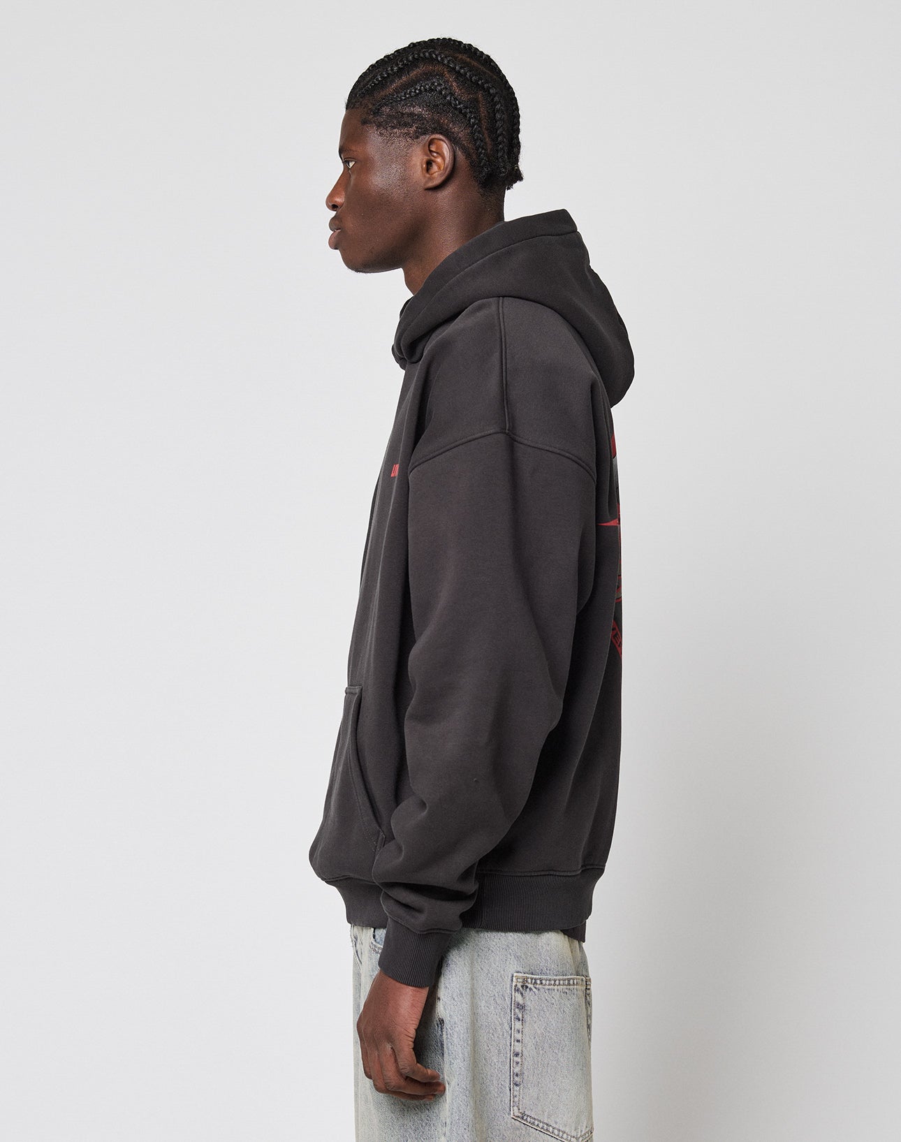 A person with braided hair stands sideways, wearing a black LFDY Globe Hooded by LIVE FAST DIE YOUNG and light blue jeans against a plain, light background.