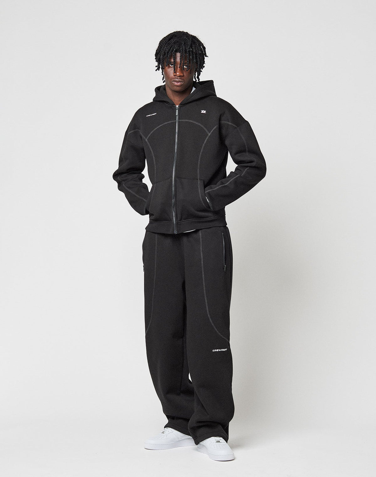 A person stands against a white background wearing the LIVE FAST DIE YOUNG LFDY Active Fleece Hooded Zip in black with matching black pants featuring white stitching, and white sneakers. Their hands are in the jacket pockets.