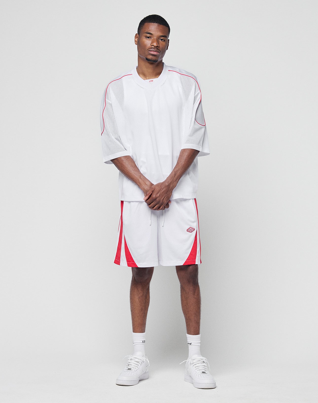 A man faces the camera against a plain background, wearing LIVE FAST DIE YOUNG Striped Mesh Shorts and a loose white athletic shirt with red accents, subtle logo, white socks, and sneakers. The regular-fit polyester outfit completes his relaxed look.